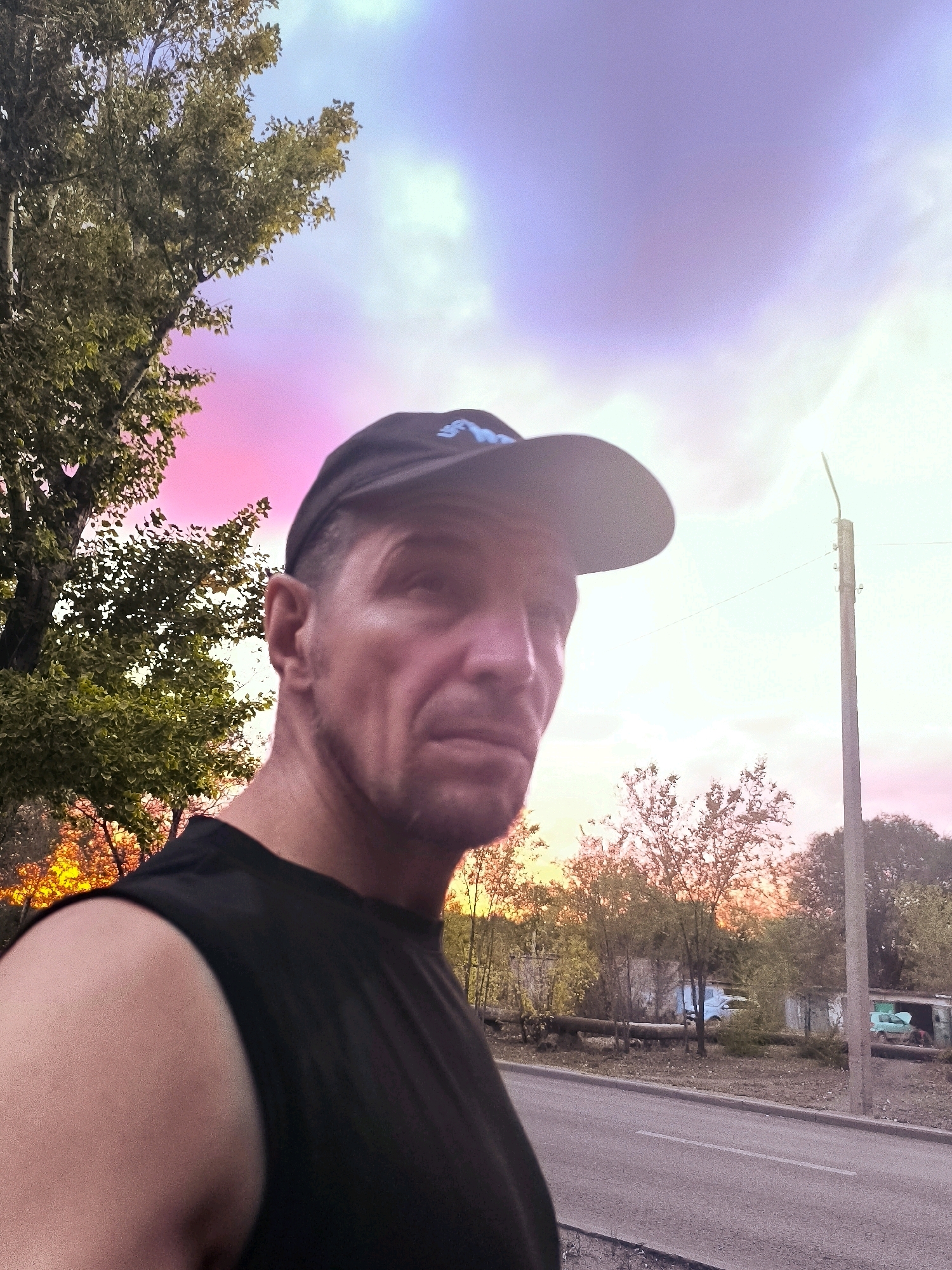 In the city of Qarağandı, Kazakhstan, a man wearing a black t-shirt and a baseball cap is captured in a close-up, self-taken photograph. The man is positioned on the right side of the frame, with his face turned towards the camera but his gaze slightly averted. He has a serious expression. The background features a road, trees, and what appears to be a row of garages or sheds, suggesting an urban or semi-urban environment. The sky above is a prominent element, displaying a dramatic gradient of colors, transitioning from vibrant pinks and purples near the horizon to lighter blues and whites higher up, indicative of a sunset or sunrise. A utility pole stands tall on the right side of the image. The overall scene is outdoors, during twilight.