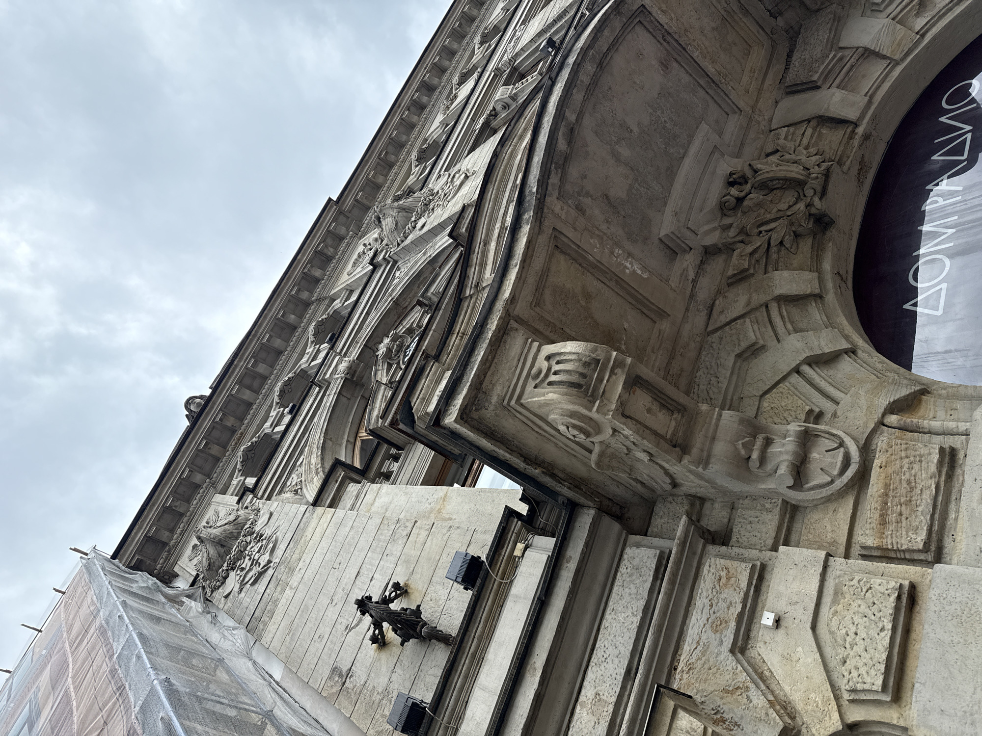The image captures a low-angle view of the facade of a historical building in Saint Petersburg, Russia. The building exhibits ornate architectural details, including intricate stone carvings, a balcony supported by decorative brackets, and a prominent cornice. Some sections of the facade are covered in scaffolding and protective sheeting, suggesting ongoing renovation or construction work. Two black outdoor lighting fixtures are attached to the wall. The sky above is overcast with a muted, grey tone. Below the balcony, a dark-colored window or display area with text is partially visible, with the Cyrillic letters "ДОМ РАДИО" discernible.