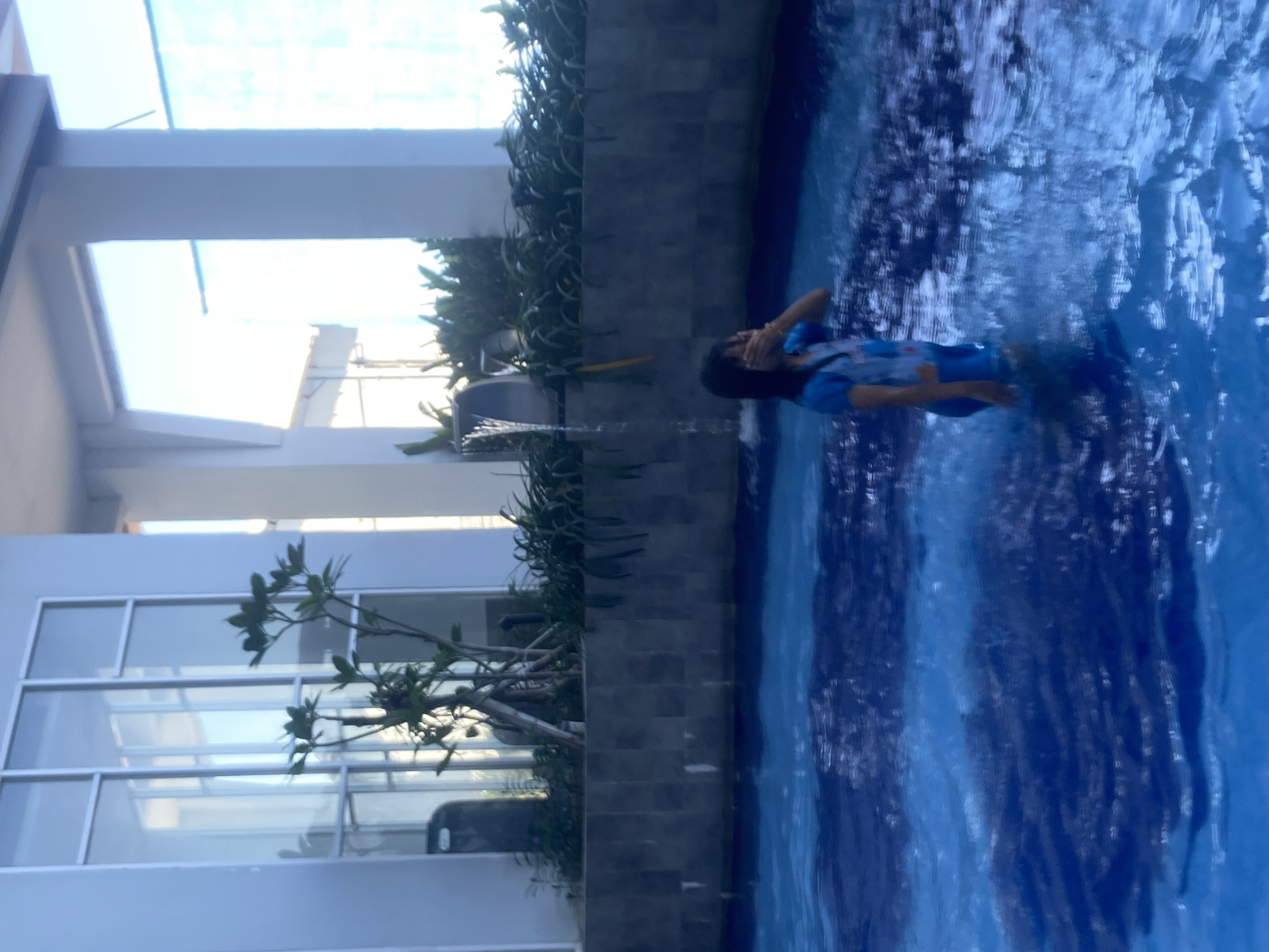 A young person wearing a blue swimsuit stands in a swimming pool, partially obscuring their face with their hand. The pool has deep blue water with visible ripples. In the background, a tiled wall separates the pool from an area with plants and a small waterfall feature. Beyond that, a building with large windows and columns is visible. The scene appears to be an indoor or covered swimming area.