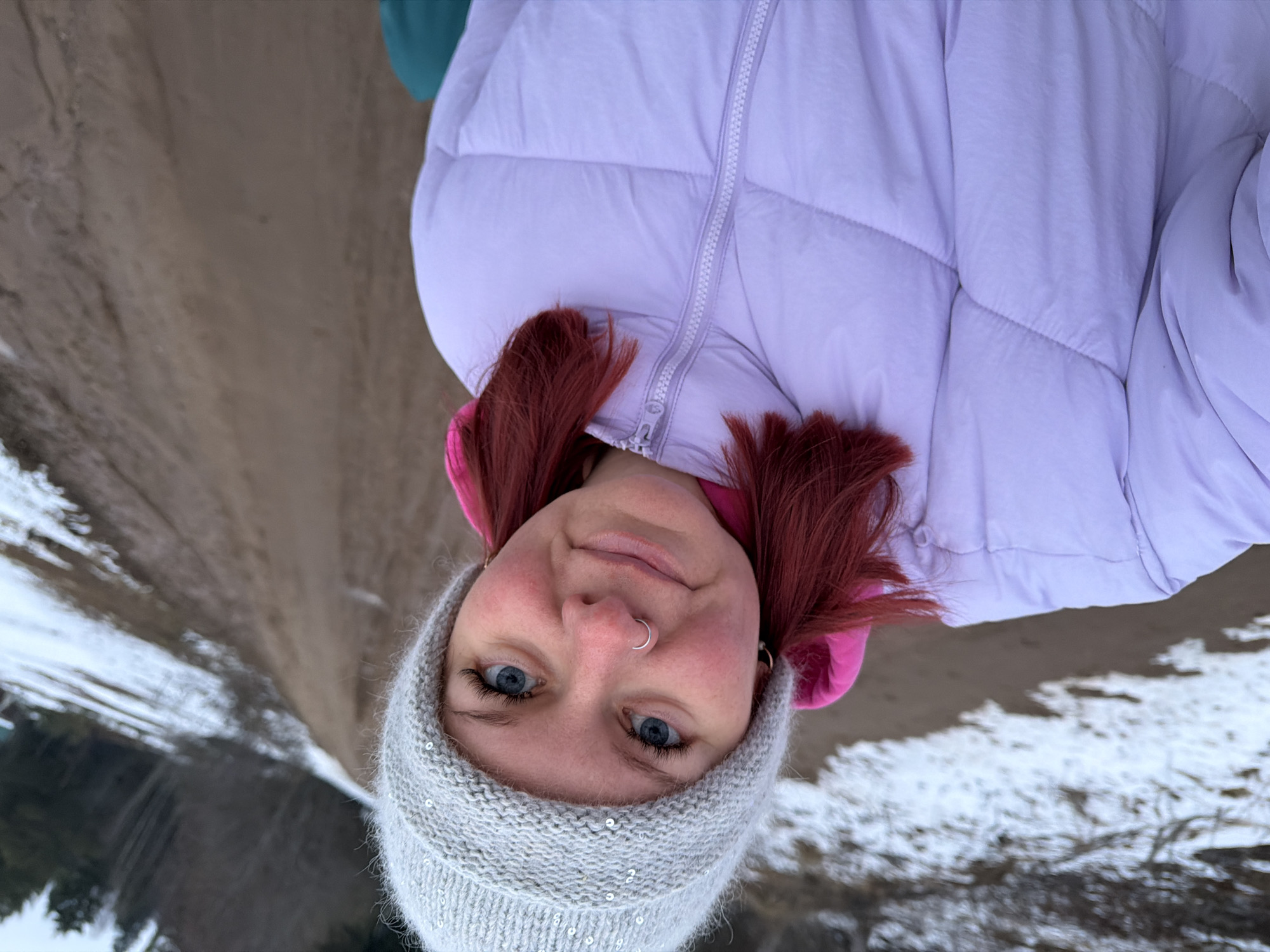 A woman with blue eyes and red hair is outdoors wearing a gray knit hat with sequins, a lavender puffer jacket with a pink hood, and a silver nose ring. The background shows a landscape with snow, sand, water, and trees. This image was taken at Chaikovsky, Russia.