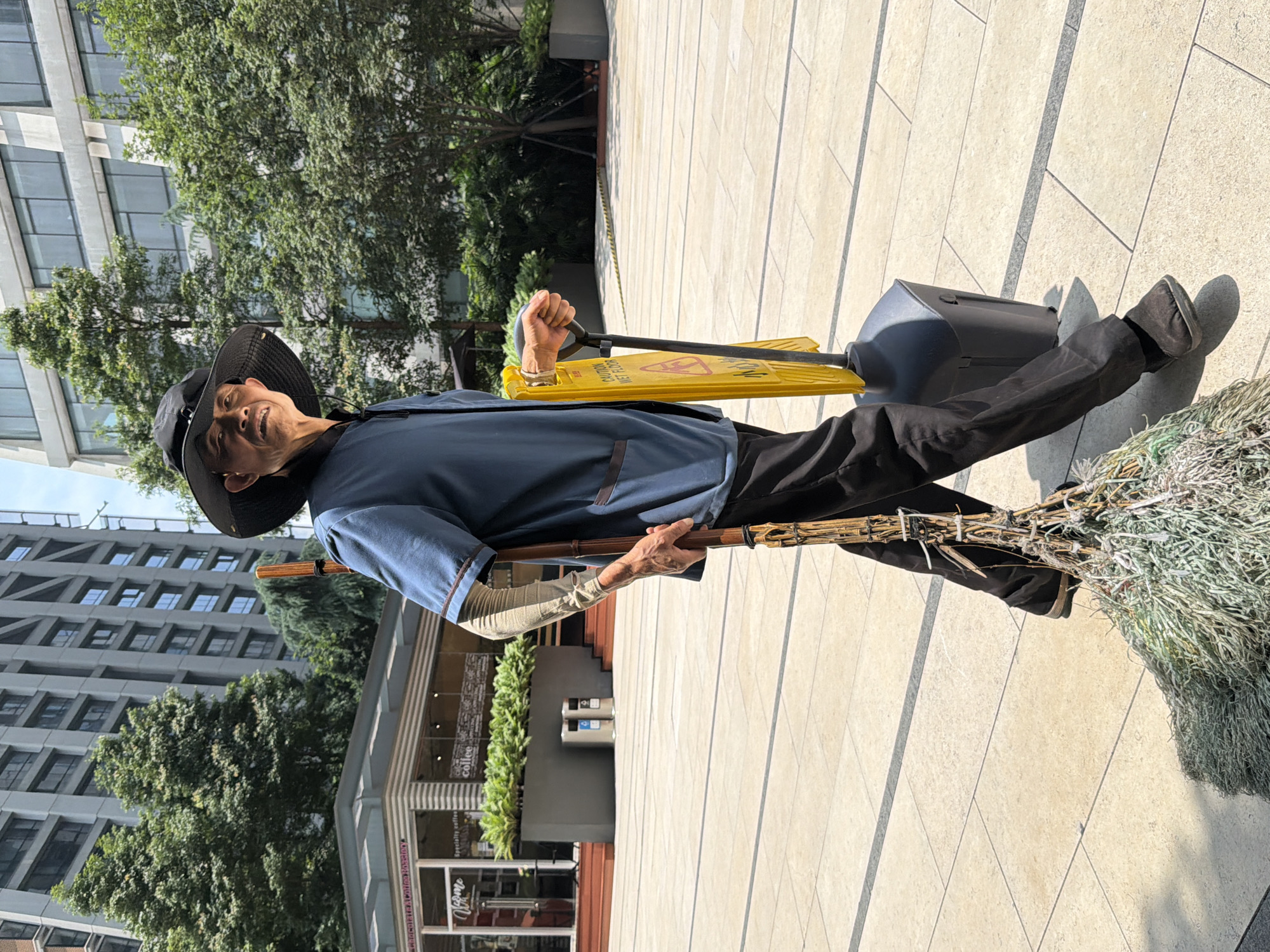 An elderly man wearing a wide-brimmed hat and a blue uniform walks outdoors on a sunny day in Yuzhong District, China. He is carrying a traditional broom made of natural materials in his right hand and holding a yellow "caution wet floor" sign attached to a black base with his left hand. His left arm is covered by a beige sleeve. He is wearing dark pants and dark shoes. Behind him, there are modern buildings and lush green trees, indicating an urban setting. The ground is paved with large, light-colored tiles. A building with glass windows and a storefront with signage is visible in the background to the left.