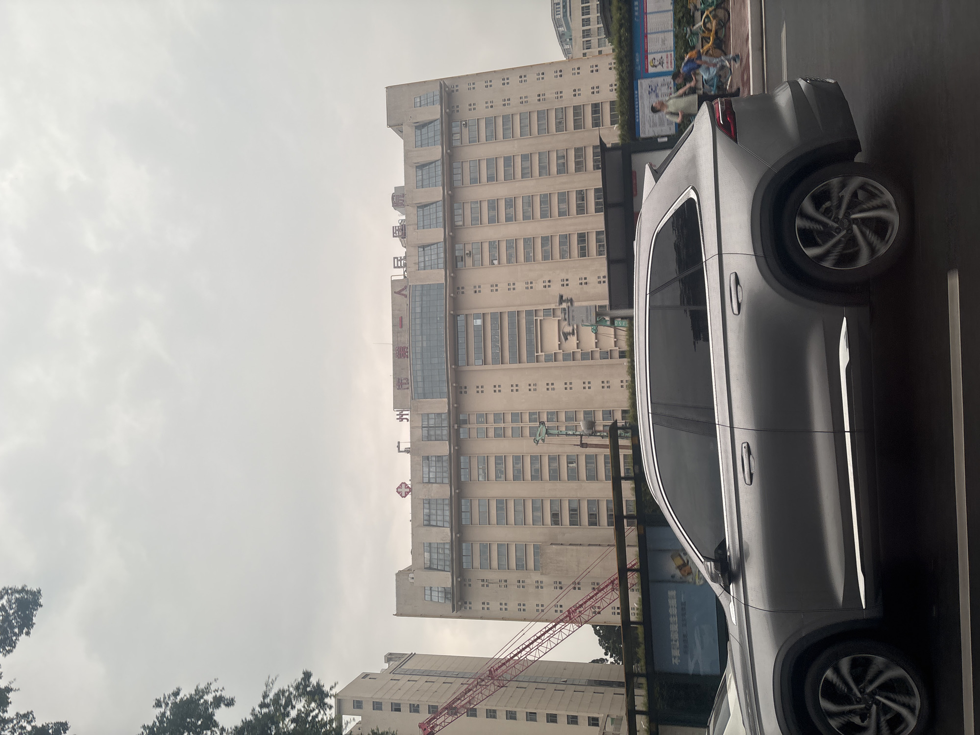 The image depicts a cloudy daytime scene in Yuexiu District, China, focusing on a large, light-colored, multi-story building with numerous windows. The building has a red emblem at its peak, along with Chinese characters that read "人民医院" (People's Hospital). To the left of this building, another taller, light-colored building is partially visible, and a large red construction crane extends upwards and across the frame. In the foreground, a silver SUV is driving on a street. To the right of the SUV, a sidewalk area is visible with a few people and bicycles. One person appears to be walking, and two children are present, one of whom is on a bicycle. Behind the people and bicycles are various posters and signs. The overall environment suggests an urban setting with ongoing construction and pedestrian activity.