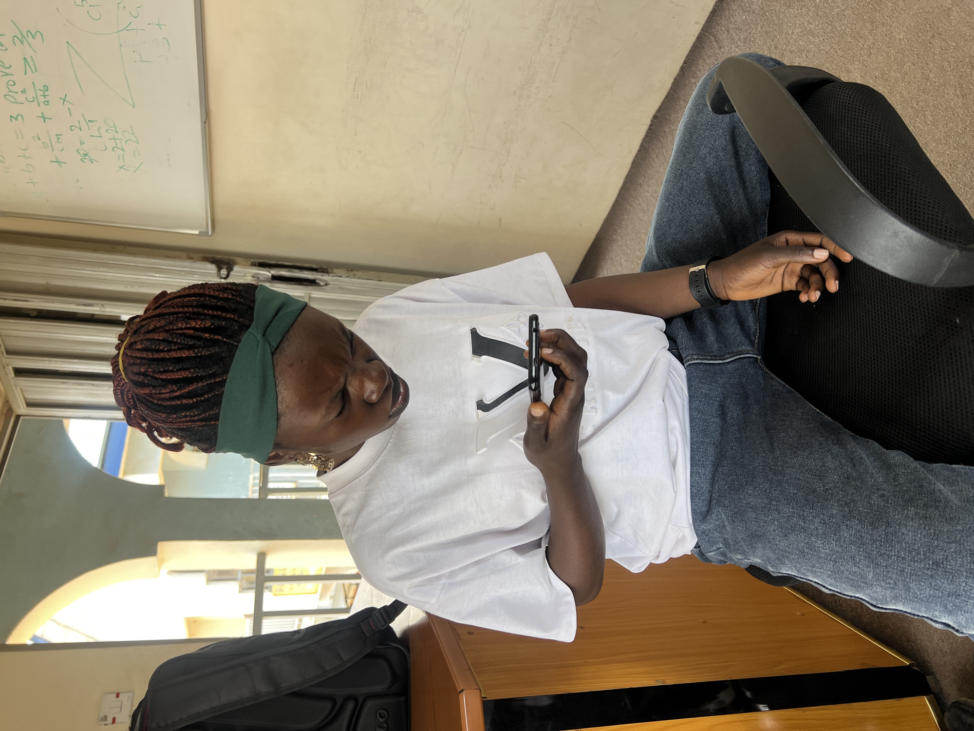 A young Black woman with braided hair and a green headband is sitting on a black office chair, looking down at her phone. She is wearing a white t-shirt and blue jeans. She is holding the phone in her right hand. A black backpack is visible on a wooden desk to her left. In the background, a whiteboard with mathematical equations is mounted on the wall. The scene appears to be an indoor location, possibly a classroom or office, in Jos, Nigeria.
