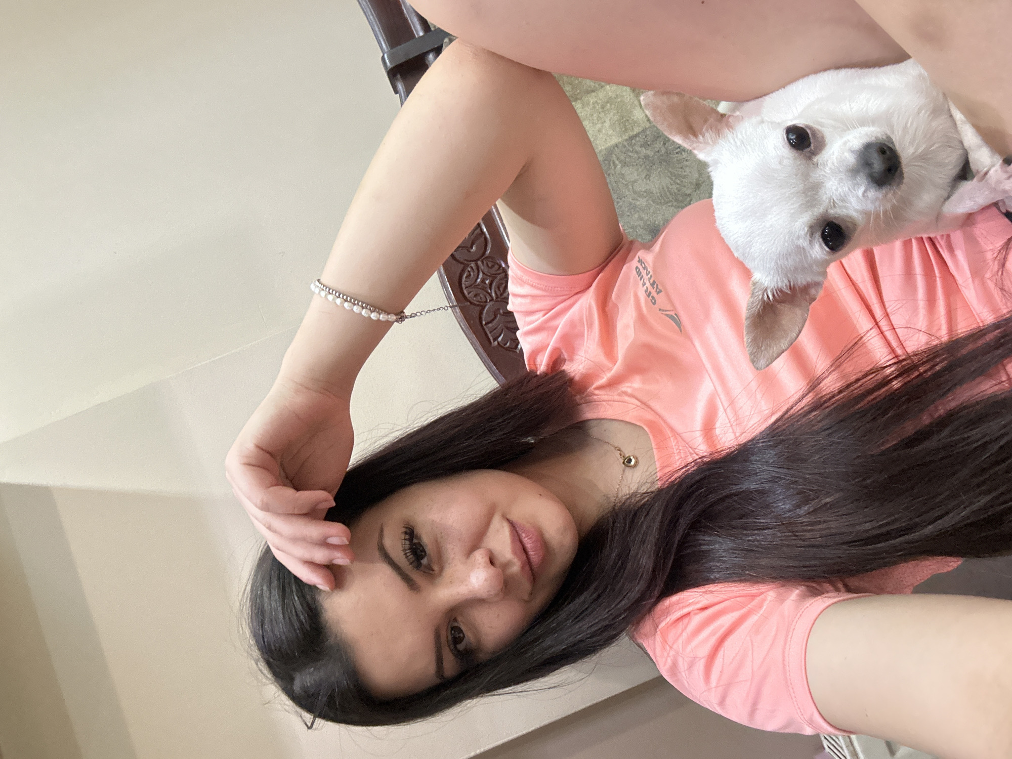 This is a selfie-style photograph taken indoors, showing a young woman with long, dark hair and a white Chihuahua. The woman is wearing a coral-colored t-shirt and a pearl bracelet. She is holding her hand to her forehead. The dog is looking at the camera. The background appears to be a room with beige walls and wooden furniture. The photograph does not provide any visual information to indicate the location in the city of Sliven, Bulgaria.