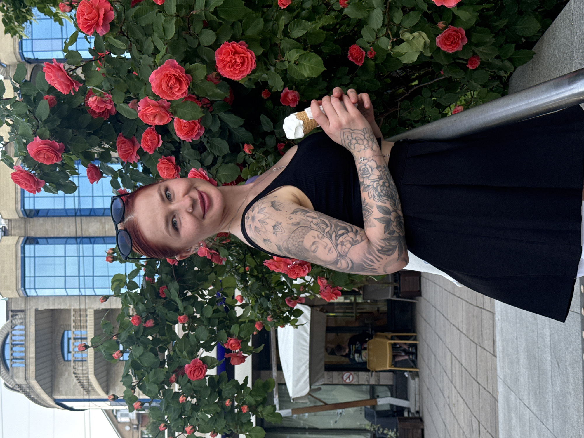 A young woman stands beside a flowering rose bush, holding an ice cream cone. She is fair-skinned with reddish hair and extensive tattoos covering her arms. She wears a black sleeveless top and a black skirt with a white underskirt visible at the hem. Sunglasses are perched on top of her head. The rose bush is full of red roses and lush green foliage. In the background, there is a building with blue glass windows and a street scene with an awning and outdoor seating. The location is Rostov-on-Don, Russia.