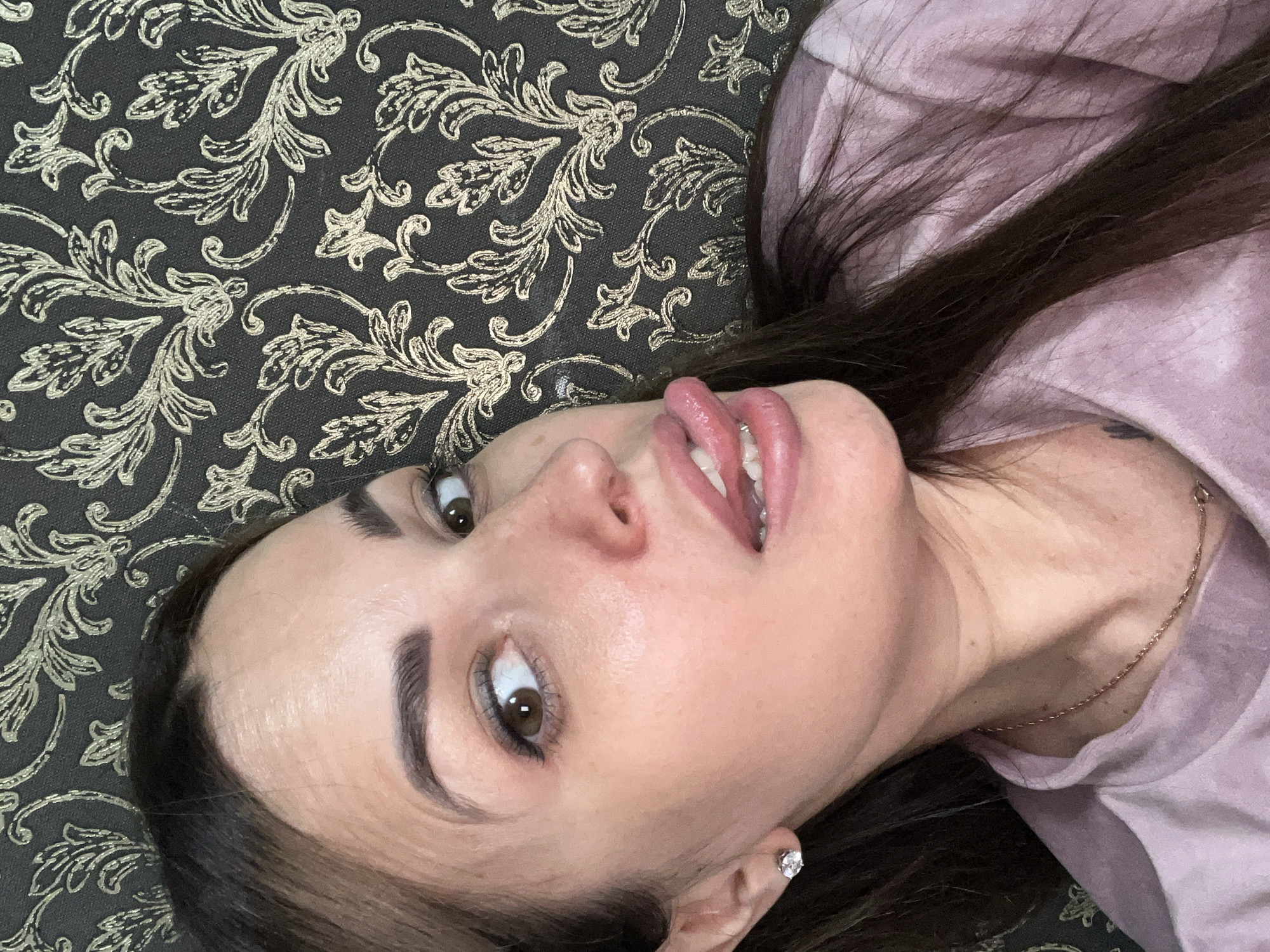 A close-up selfie shows a woman with brown hair and brown eyes. She is wearing a pink shirt and a gold necklace. Her tongue is sticking out and she is wearing a diamond earring. She is against a dark patterned background.