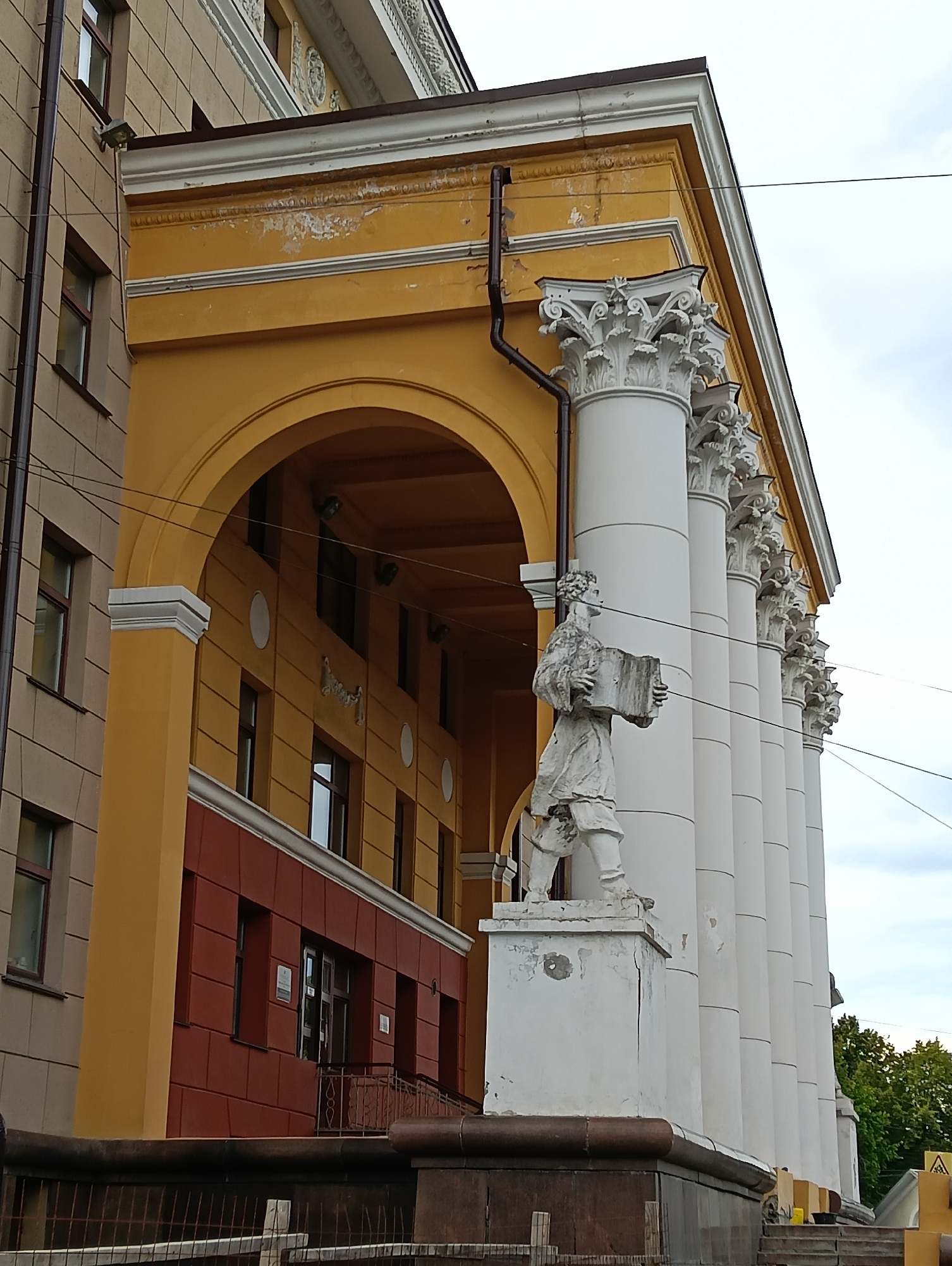 The image shows the facade of a building in Petrozavodsk, Russia, specifically the building of the National Library of Karelia. A statue of a man holding a book stands in front of a colonnade of white pillars. The building has a yellow arched portico on the left, and a red section with windows below it. The sky is overcast.