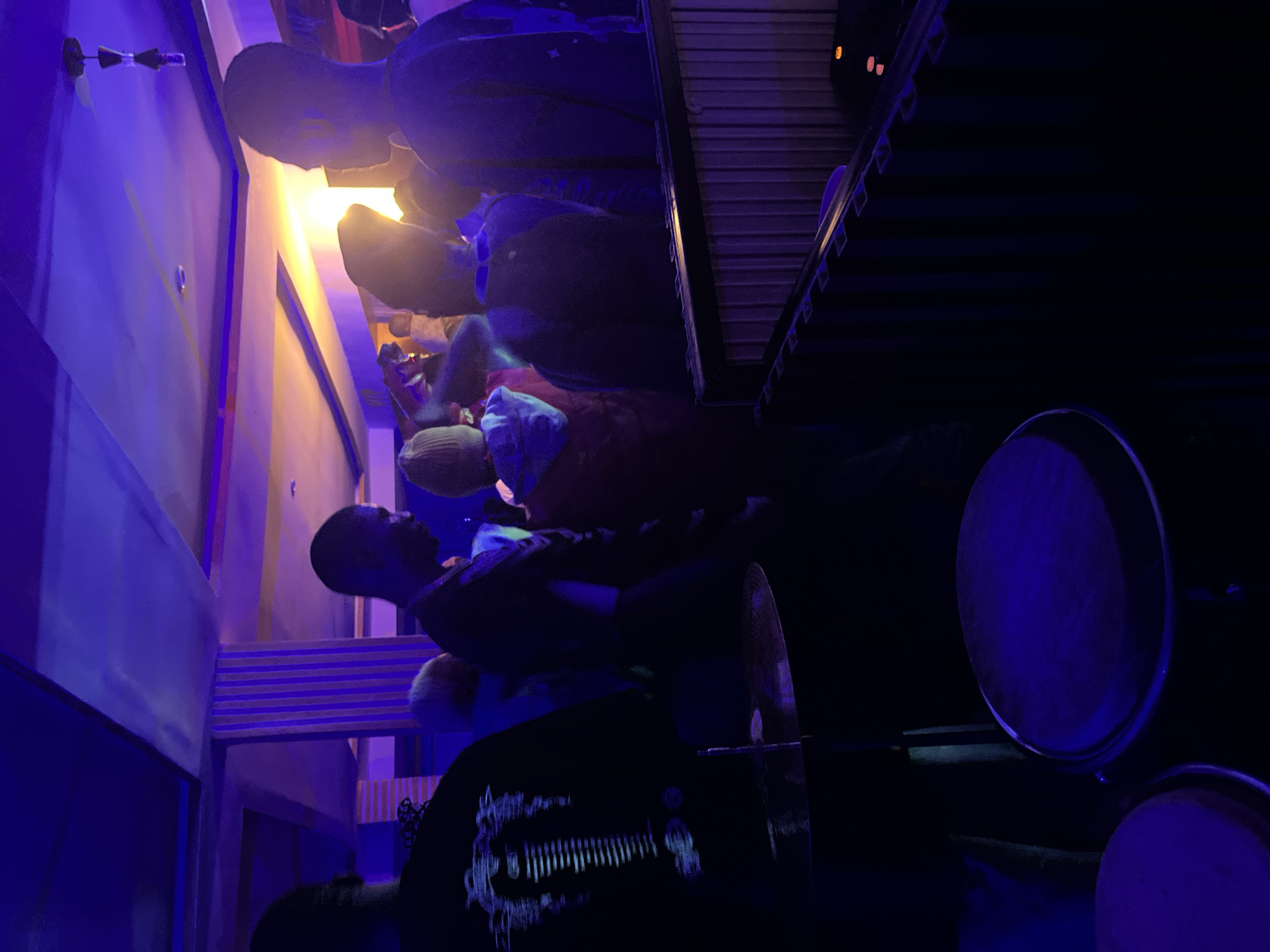 The image depicts an indoor scene, likely a club or performance venue, with a lively atmosphere illuminated by blue and yellow lighting. Several individuals are visible, some facing away from the camera, suggesting they are engaged with a performance or social activity. In the foreground, a drum and cymbal are set up, indicating a musical performance is in progress or about to begin. The environment appears to be a public space with decorative pillars and ceiling elements. The specific location in Wumat, Nigeria, is not evident from the visual information provided.