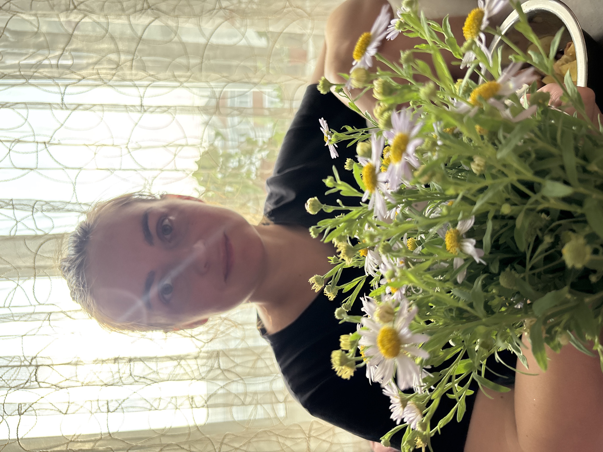 A woman with light brown hair pulled back in a bun sits behind a bouquet of white and yellow wildflowers. She is wearing a black t-shirt. The background is a sheer, patterned curtain with sunlight streaming through it. A sliver of a building and trees is visible through the curtain. The scene is likely indoors, possibly a home, in Blagoveshchensk, Russia.