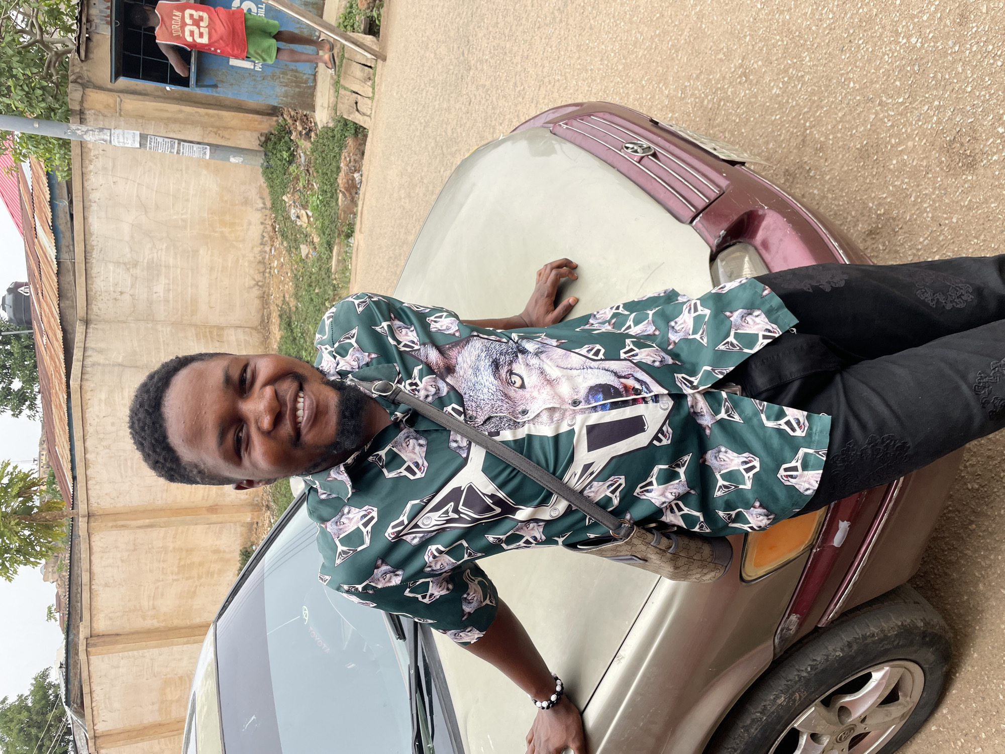A young Black man with a beard and short, dark curly hair smiles broadly at the camera, leaning on the hood of a light gold sedan. He wears a dark green collared shirt adorned with a repeating pattern of wolf heads and geometric shapes, black trousers, and a black and white beaded bracelet on his right wrist. A dark strap, possibly from a small bag, crosses his chest.
The scene takes place outdoors, likely on a street or unpaved area in Jos, Nigeria, during daytime under an overcast sky. The ground beneath the car is a textured mix of dirt and small stones.
In the background, a person in a red basketball jersey with "JORDAN 23" on the back and green shorts stands in a doorway or window opening of a light-colored concrete building. Behind this person, a blue structure with faintly visible text reading "PAY BILL" is present. To the left, a building with a corrugated metal roof is visible, and a utility pole stands prominently with some illegible posters attached.
The car, which appears to be a Toyota sedan based on a faint logo on the windshield, has a reddish-brown or maroon front grille/bumper section and shows signs of wear, including a minor scratch near the front right headlight. The man appears relaxed and happy, posing for the photo.