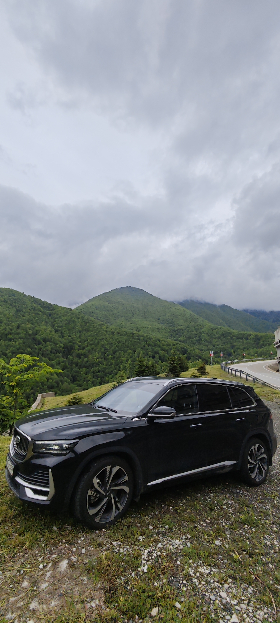 A black GAC GS8 SUV sits on a dirt and grassy shoulder near a mountain road in Chvizhepse, Russia. The mountains behind the car are covered in green forest under an overcast sky. Roadside guardrails and road signs are visible in the background, indicating a winding road. The car faces toward the left and is angled slightly away from the viewer.