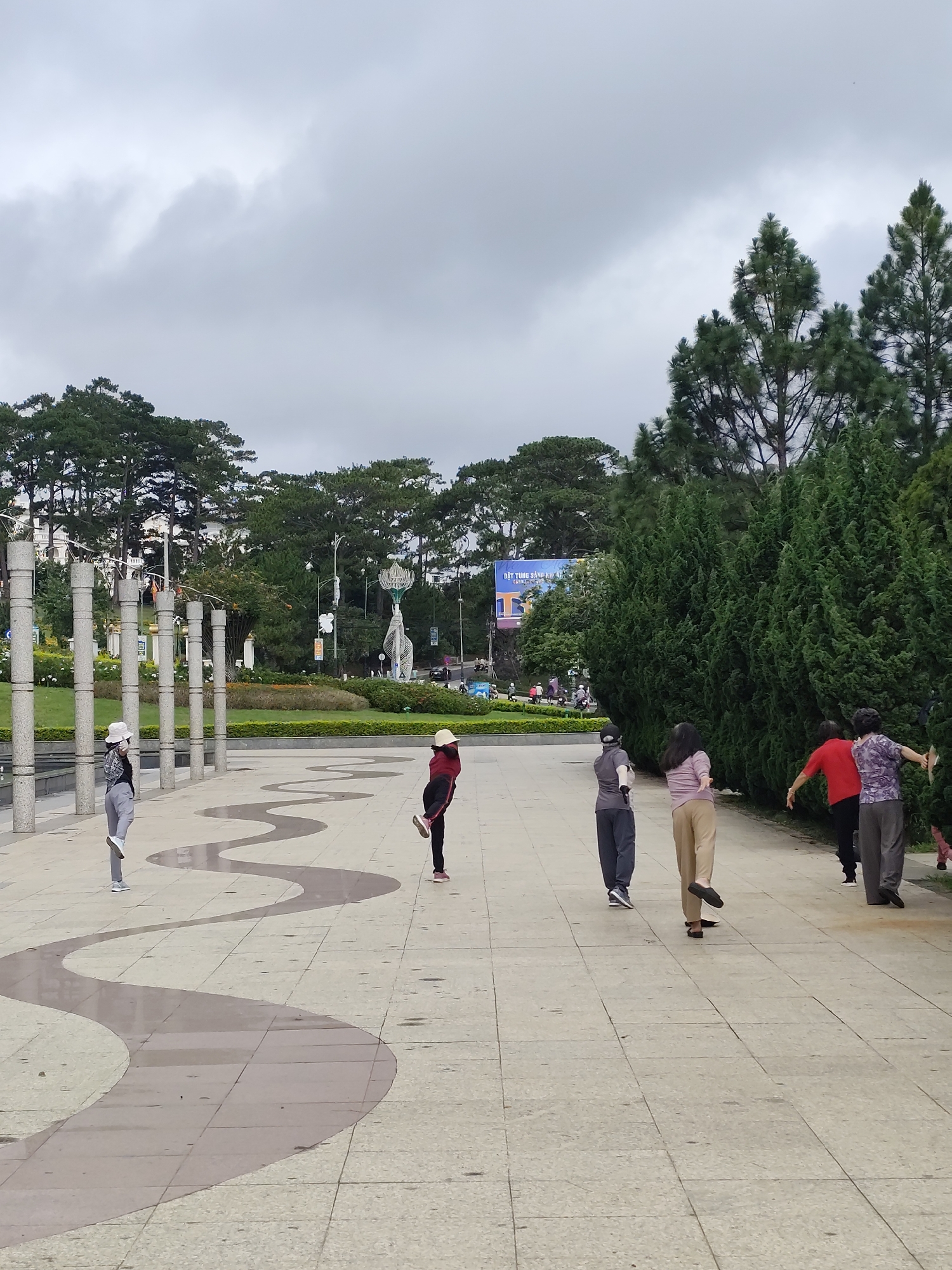 The image captures a scene in Da Lat, Vietnam, featuring a group of six women engaged in a form of exercise or dance on a paved plaza. The plaza is characterized by a light-colored tiled surface with a darker, serpentine pattern running across it. To the left, a row of cylindrical concrete columns adds a structural element to the space, beyond which is a grassy park area, with trees, decorative lighting, and a sculpture. In the background, a billboard is visible. The group of women are spaced apart and in mid-motion, with one leg lifted off the ground. They are wearing casual attire and face masks. The right side of the image is dominated by dense, evergreen trees. The sky is overcast, suggesting a cloudy day.