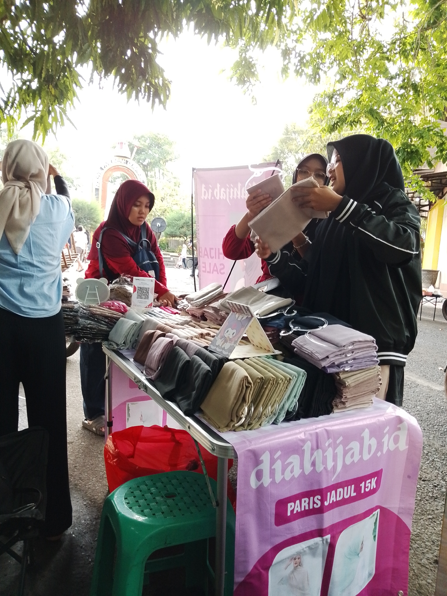 At a vendor's stall in Kecamatan Pati, Indonesia, three women are engaged in a transaction involving textiles, likely hijabs. A woman with a blue hijab is standing to the left of the stall, her back to the camera. A woman in a maroon hijab stands behind the vendor table examining an item. Two women, one wearing a black hijab with white stripes and the other a lighter hijab, are closely inspecting a tan hijab. The stall is covered with a pink cloth bearing the text "diahijab.id" and "PARIS JADUL 15K." A wide variety of hijabs are stacked on the table, along with a small stand with an image of a cartoon character. Underneath the table, there is a green plastic stool and a red bag. The environment includes an arched entrance to a park in the background and foliage overhead, suggesting an outdoor location.