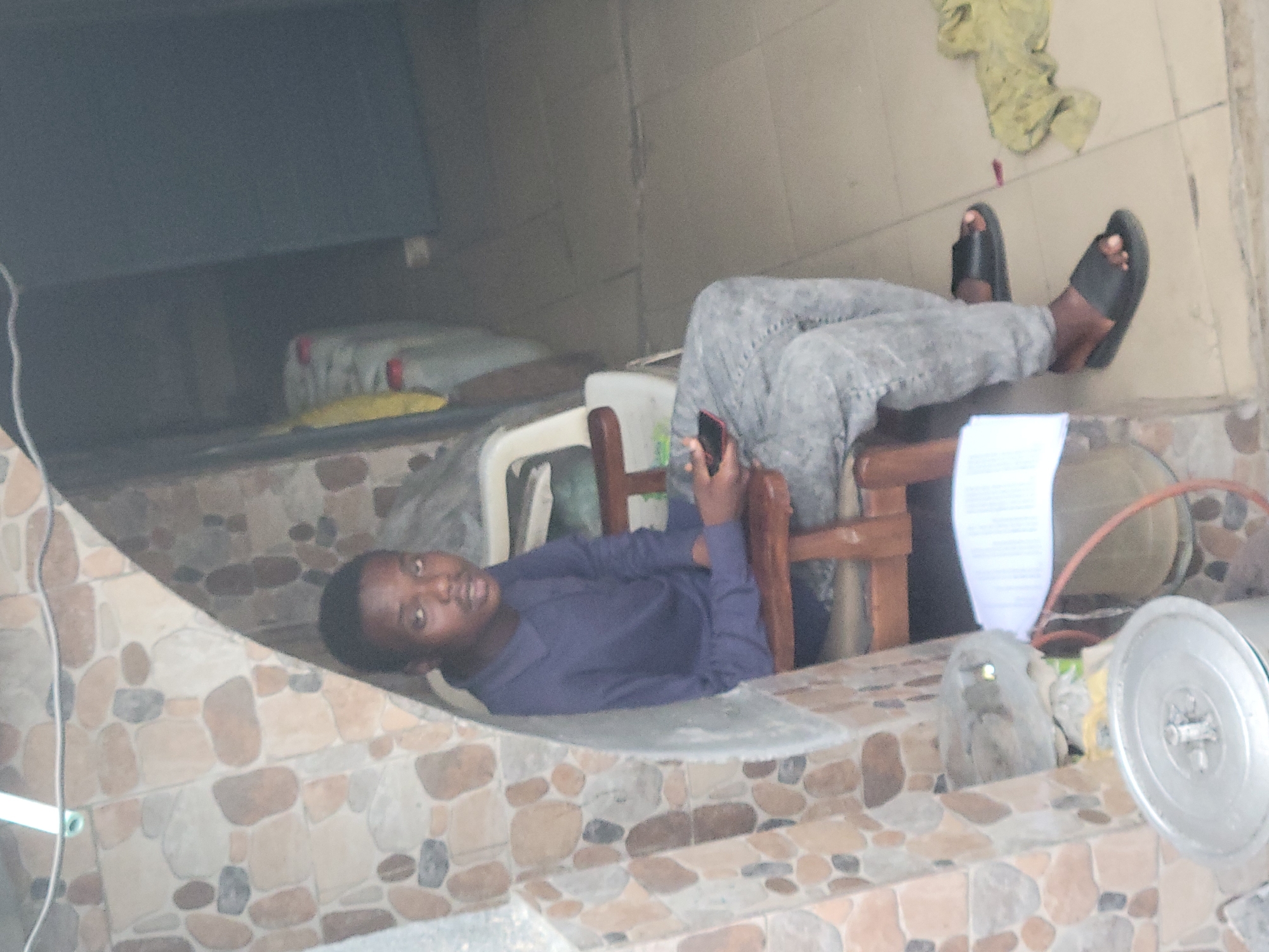 A young man in Jos, Nigeria, is lying on a tiled surface with his legs resting on a chair. He is wearing gray pants and black sandals. He is holding a cell phone in his right hand. Beside him is a wooden chair and a metal object. The background consists of tiled walls and a shelf-like surface above him.
