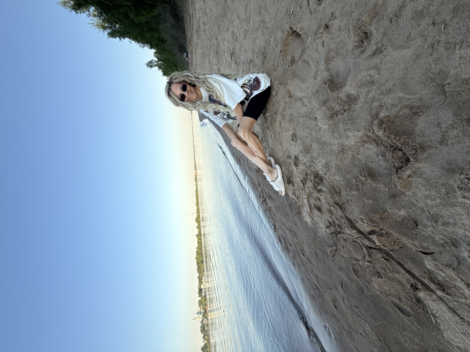 A person wearing a white shirt, black shorts, and white sandals stands on the sandy shore of a body of water. The person has long, blonde hair and is wearing sunglasses. The water is calm, and the sky is clear blue. In the distance, buildings and trees are visible along the opposite shoreline. The location is identified as the city of Rusanovo, Russia.