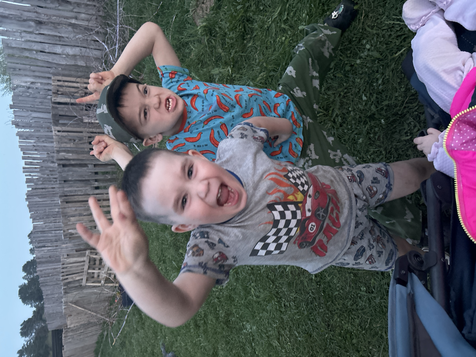 Two young boys are lying on green grass in an outdoor setting. One boy wears a blue shirt with a chili pepper pattern and a green cap. He is making a hand gesture, possibly devil horns. The other boy, closer to the camera, wears a grey Cars-themed pajama set and has his mouth open in a wide smile while holding up his hand with fingers splayed.
A partial view of a stroller is visible on the right side of the image, with a pink and white garment near it. In the background, there is a wooden fence. The location is Rusalevka, Russia.