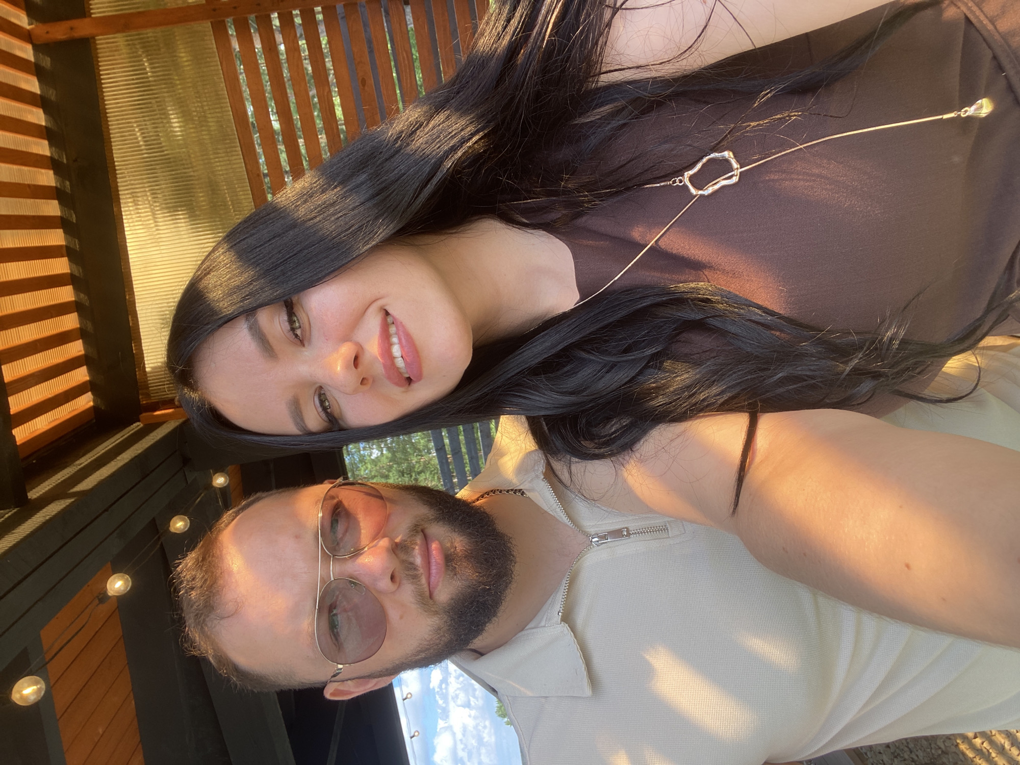 A couple takes a selfie outdoors, under a wooden gazebo with string lights. The man, on the left, has short brown hair and a beard, wearing sunglasses and a light-colored zip-up polo shirt. The woman, on the right, has long dark hair and is wearing a brown dress with a long pendant necklace. The sunlight casts a warm glow on them, and the background shows trees and a fence, suggesting a location in a rural or suburban area, possibly the city of Chaikovsky, Russia, given the context.