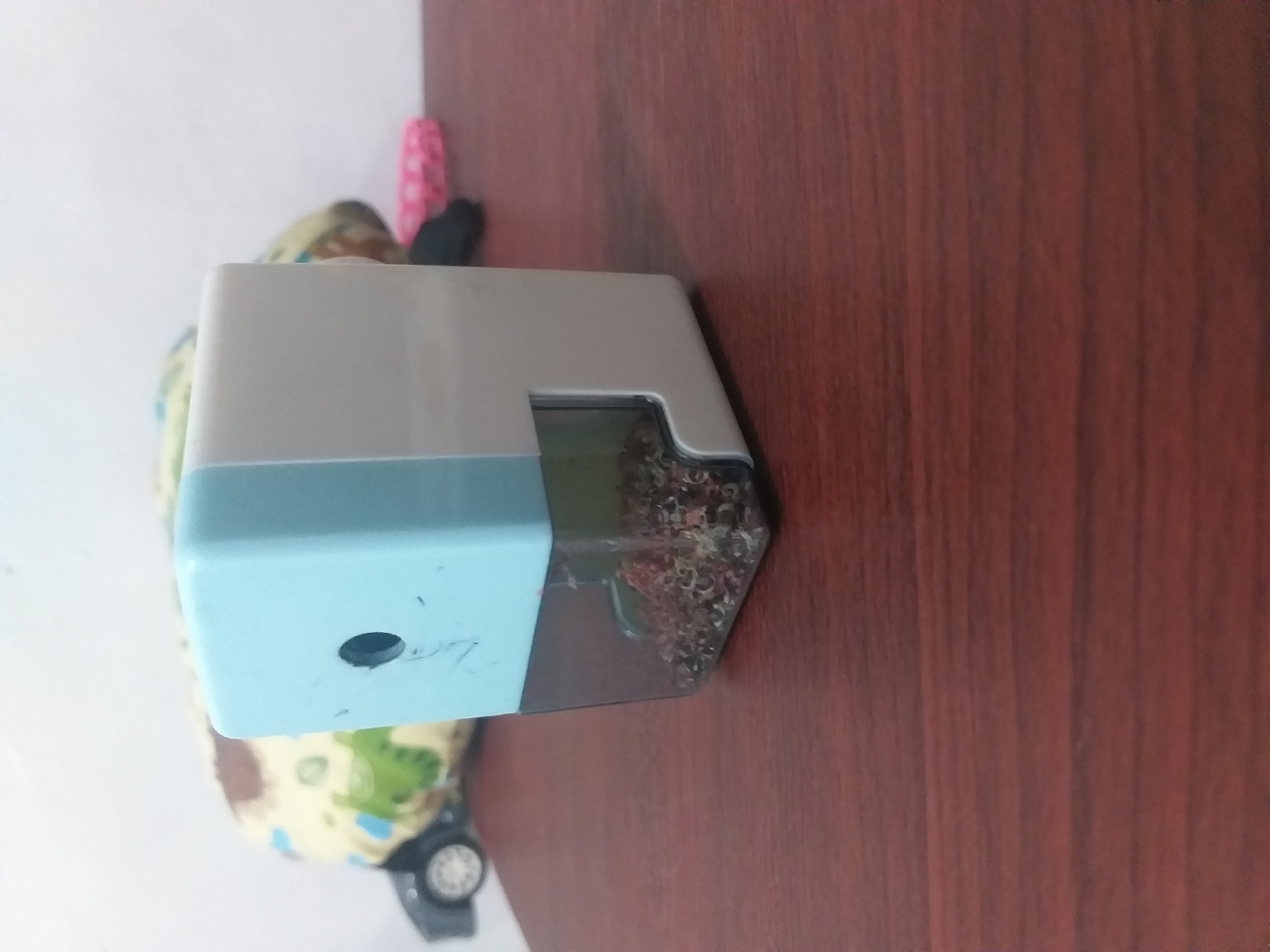An overhead shot shows a light blue and white pencil sharpener filled with pencil shavings, sitting on a dark brown wooden surface. In the background, out of focus, is a soft toy with a green and brown pattern and a small black toy car. There is also a blurred pink object. The scene appears to be indoors.
