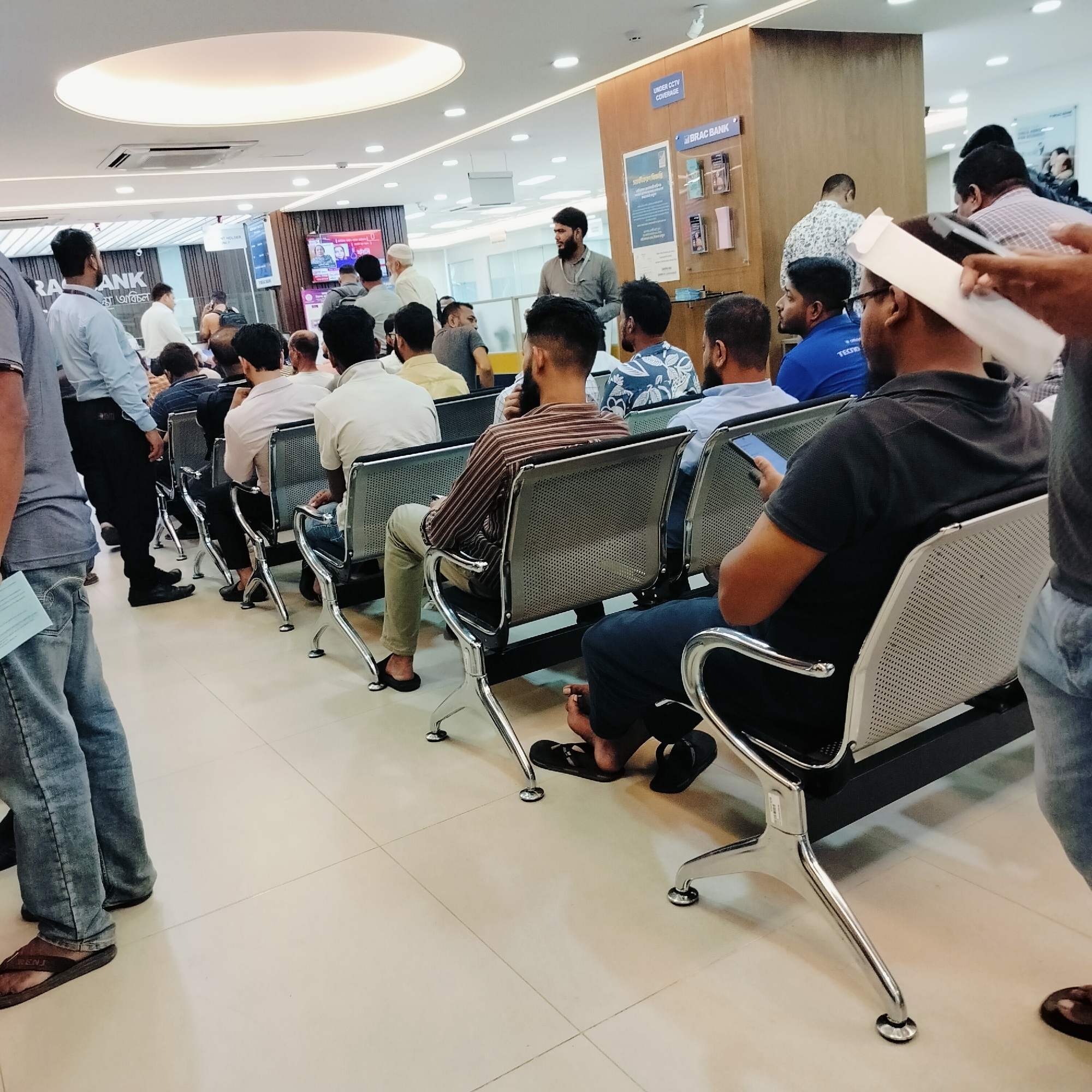 The image captures the interior of a BRAC Bank branch, serving as a customer waiting area, located in an unknown city and region. Approximately 20-30 individuals, predominantly adult males, occupy the space. Most are seated in multiple rows of modern, connected metal chairs featuring perforated backs and seats, with polished chrome frames, arranged on a light-colored tiled floor.
Bright overhead lighting is provided by numerous recessed circular fixtures and a large, central oval-shaped luminous ceiling panel. An air conditioning vent is also visible on the ceiling. The walls are a mix of light-painted surfaces and sections paneled with dark wood.
In the left foreground, a man in a blue t-shirt and light-wash jeans, wearing brown flip-flops, stands and holds a light blue paper in his left hand. In the right foreground, a man in a dark grey polo shirt and dark trousers is seated, engrossed in a smartphone held in his right hand; his bare feet rest on dark flip-flops beside him. Adjacent, another seated man wears a brown and white striped long-sleeved shirt and light olive trousers.
Mid-ground, a bearded man in a grey long-sleeved shirt stands, facing slightly right, observing the room. Further back, a man in a light blue collared shirt with an ID badge stands near a counter. A television screen mounted on a back wall displays two male figures, potentially part of a news program or bank advertisement. Prominent "BRAC BANK" branding is visible on signs and informational posters. A vertical sign on a wooden panel states "UNDER CCTV COVERAGE." The atmosphere is active, indicating a busy operational period for the bank.