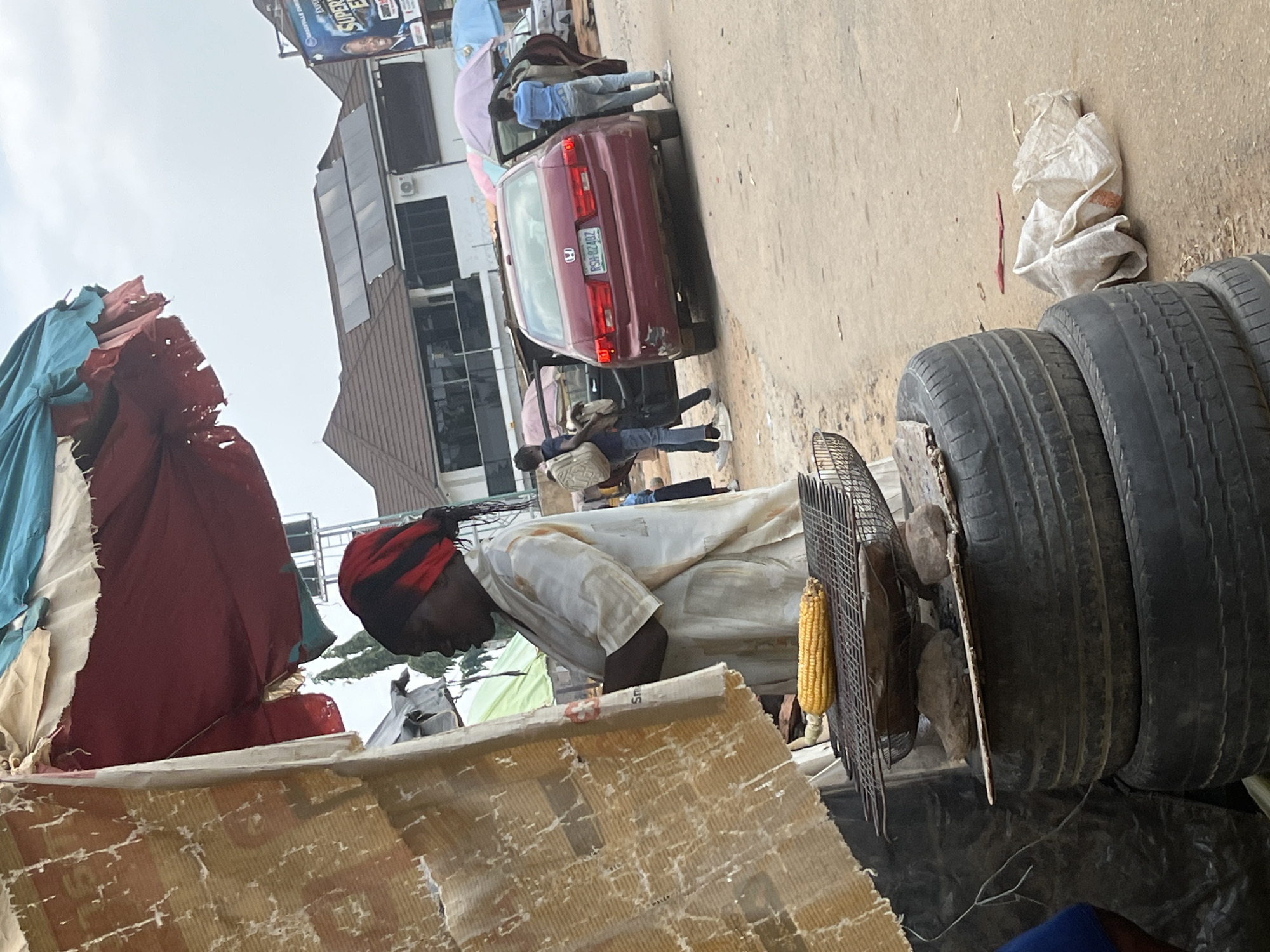 The image depicts an outdoor scene in what appears to be a market or roadside stall in Abuja, Nigeria. The primary subject is a person, likely a vendor, wearing a white garment and a red and black head covering. They are bent over, tending to a small charcoal grill where a cob of corn is visible. Beside the grill, a fan with a metal grate is positioned.
In the background, a red sedan is parked, with its doors open. Two other individuals are seen near the car. The setting includes various objects like stacked tires, bags, and tarps or fabric draped over structures, suggesting a makeshift stall or storage area. The ground appears to be a paved surface, typical of a street or marketplace.
The lighting suggests it is daytime, possibly overcast. The overall impression is one of everyday commerce and activity in a busy, possibly informal, urban environment. There is no easily discernible text visible in the image, apart from a license plate on the car which is too blurry to read.