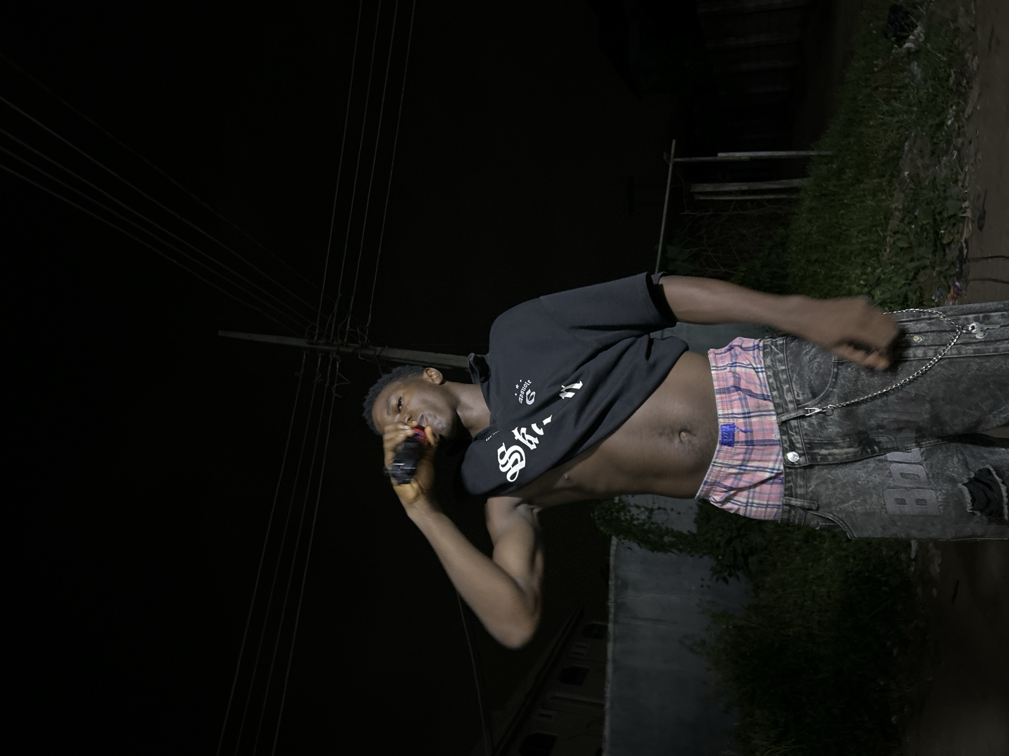 A man stands outdoors in Ihiagwa, Nigeria at night, wearing ripped black jeans with a chain, plaid boxers, and a black short-sleeve shirt pulled up to reveal his torso. He holds a black object to his mouth. Overhead power lines are visible against the dark sky. Green foliage and a low wall are in the background.