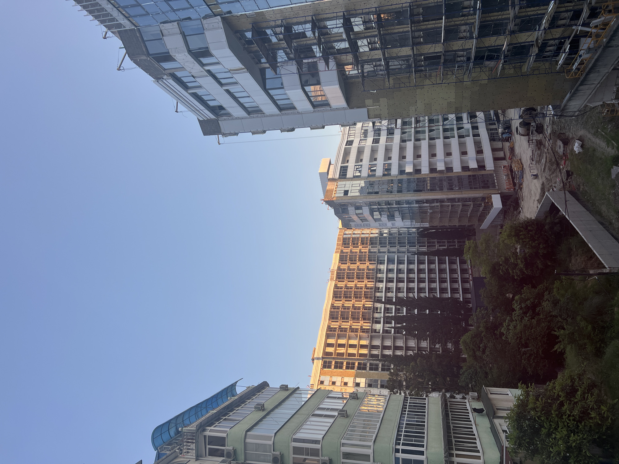 This image depicts an urban landscape in Sochi, Russia, captured under a clear, bright blue sky, indicative of morning or late afternoon due to the angle of direct sunlight.
On the left, a multi-story residential building with a light green painted facade features numerous enclosed balconies or sunrooms with white frames. A prominent translucent blue corrugated plastic canopy extends over the top-most balcony. Multiple external air conditioning units are affixed to its exterior walls.
On the right, a tall, modern-style building is undergoing construction. Its upper sections are partially clad with reflective glass panels and white facade elements, while lower sections expose scaffolding and a yellowish-green insulation material covering the structural frame. Several lifting or crane mechanisms are visible near the top edge of this building.
In the mid-ground, several other multi-story buildings are present. One large building in the center-left appears partially constructed, with its concrete frame and a grid of window openings visible. The side of this building is warmly illuminated by direct sunlight. Another building to its right is nearing completion, featuring white balconies and large windows. Further back, additional buildings contribute to the urban density.
The ground level between the buildings is an active construction site. Dirt paths, various construction materials (including large cable reels, pipes, and debris), and concrete barriers are visible. A few small figures, presumed to be construction workers, are present on the site, engaged in activity.
Lush green trees and dense bushes occupy the lower central portion of the image, separating the residential building from the construction area. Several tall, slender evergreen trees are interspersed among the buildings.