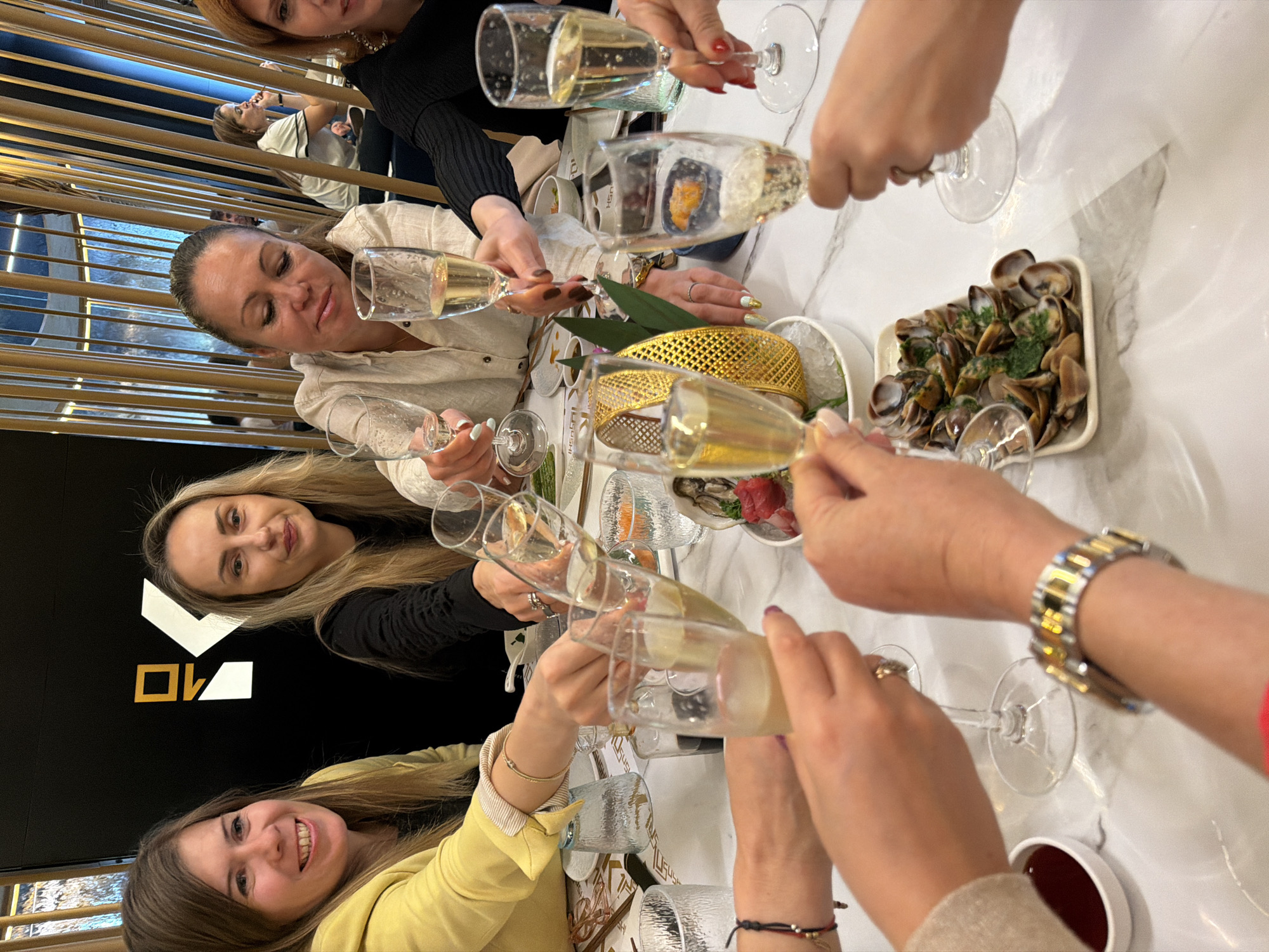 The image captures a group of women toasting with glasses of champagne at a restaurant. They are seated around a table laden with food, including sushi, seafood, and dipping sauces. The women are wearing various outfits, including a yellow jacket, a black top, and a beige blouse. One woman is wearing a notable gold and silver watch. The table has a marble top. The location is Tarragona, Spain, as it is a coastal city where this kind of seafood is common, as well as a bustling restaurant culture.