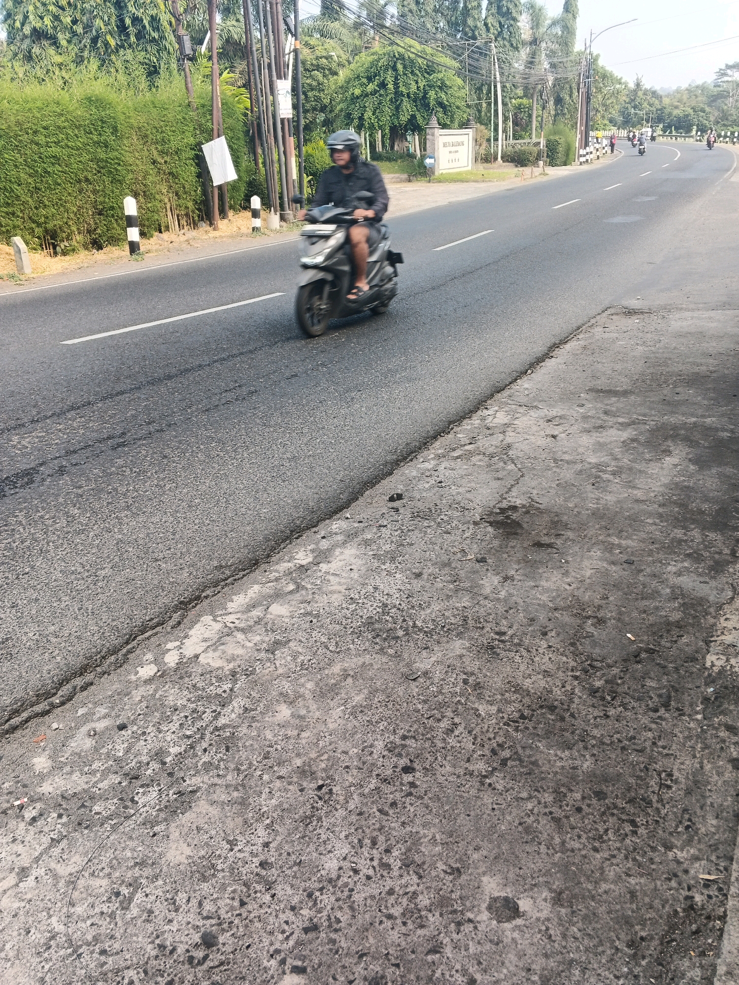 A person rides a scooter on a paved road in Ungaran, Indonesia. The scooter rider is wearing a helmet and dark clothing. The road is bordered by a dense green hedge on the left and a concrete sidewalk on the right. In the background, there are trees, utility poles, and other scooters on the road. A sign is visible on the left side of the road. The sky is bright, suggesting it is daytime.