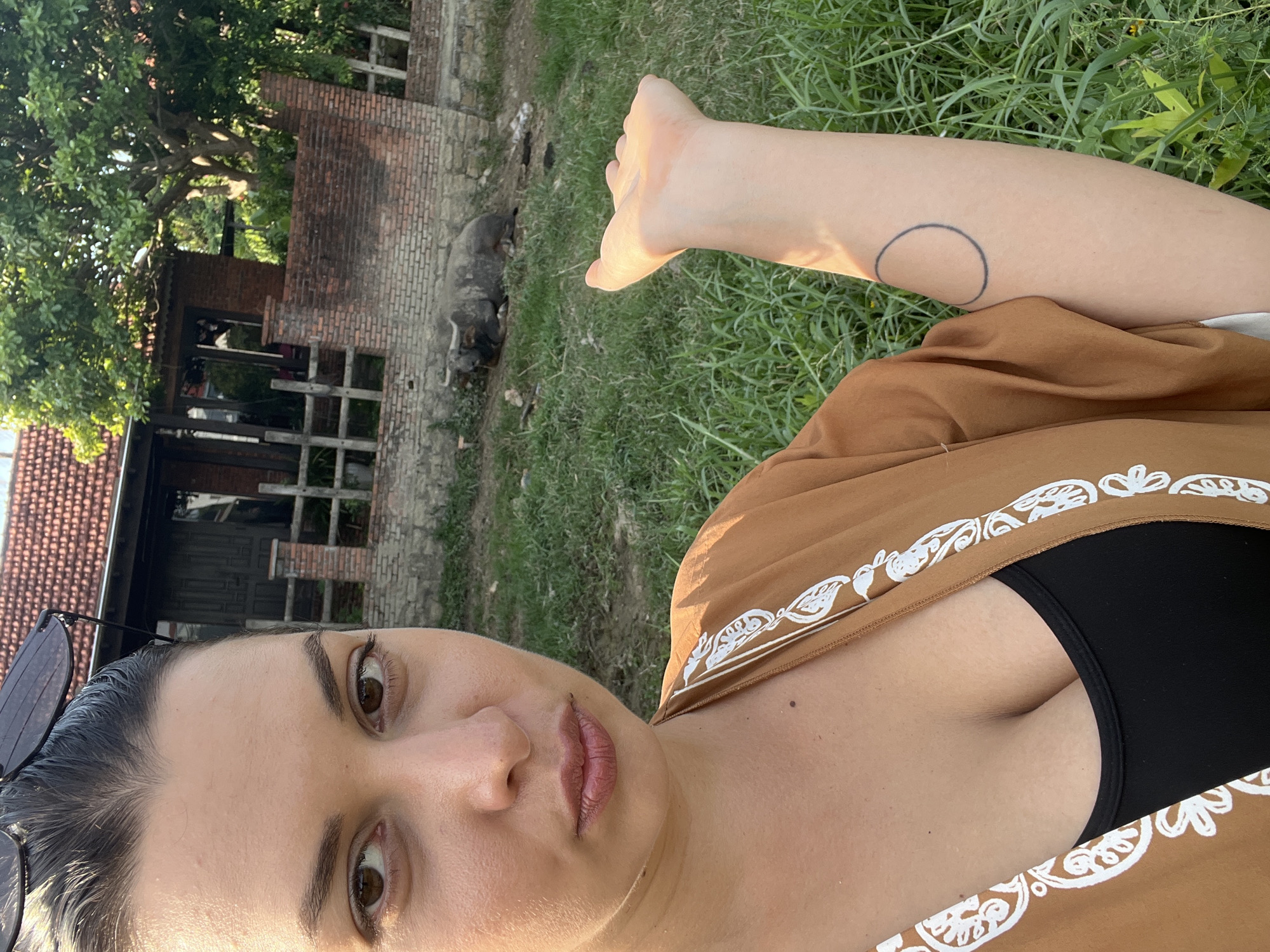 This is a selfie taken outdoors, likely in a rural setting in Đà Nẵng, Vietnam, given the presence of a water buffalo and lush greenery. The image is oriented vertically.
The primary subject is a woman, seen from the chest up, with her face close to the camera. She has dark hair pulled back, brown eyes, and is wearing a brown, patterned garment over a black top. Her expression appears neutral or perhaps contemplative. A pair of dark sunglasses rests on top of her head.
In the background, there is a partially visible brick building with a tiled roof, suggestive of a rustic structure or perhaps an older dwelling. Trees and dense green grass fill the rest of the scene. To the left of the woman, a water buffalo is lying down in the grass, an animal commonly found in Vietnamese rural landscapes.
The lighting suggests it is daytime, with sunlight casting a warm glow, particularly on the woman's arm, which is raised and shows a circular tattoo. The overall mood is relaxed and informal, capturing a moment in a natural, perhaps serene, environment. No visible text is present in the image.