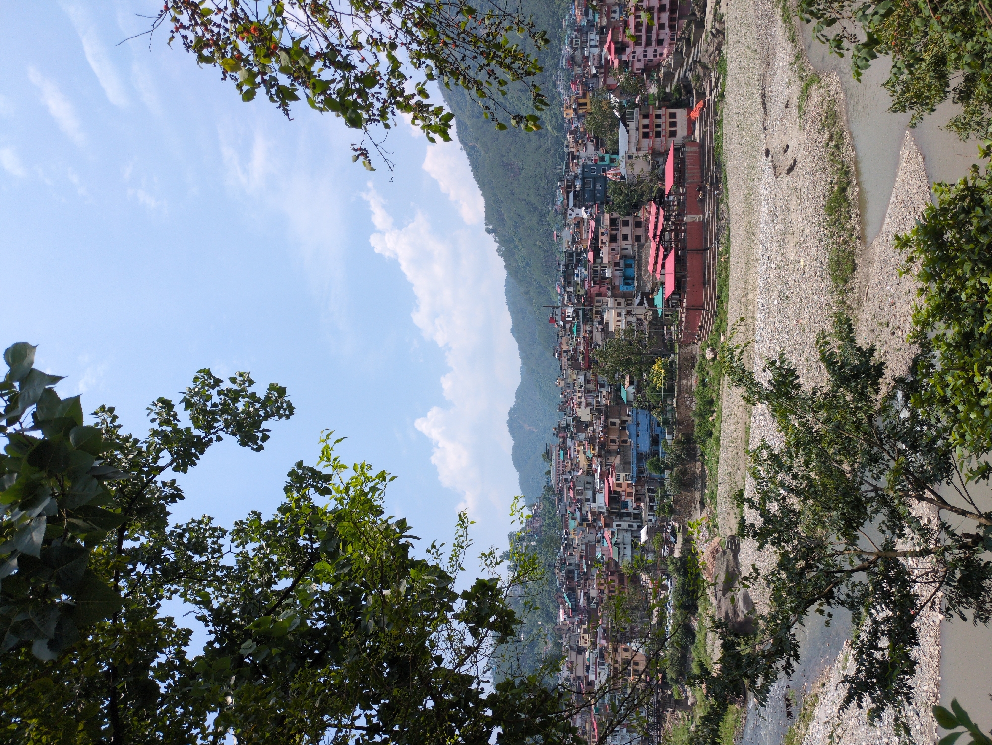 The image depicts a panoramic view of the city of Mandi, India, nestled in a valley. The city is characterized by its densely packed buildings with colorful roofs, predominantly red and pink, cascading down the hillsides. A river, appearing somewhat shallow with a rocky bed, flows through the foreground, with a bridge visible in the mid-ground. Lush green trees frame the scene from the left and right edges, suggesting a natural environment surrounding the urban landscape. The sky is a clear blue with scattered white clouds. A complex of buildings with prominent red roofs, likely religious or cultural structures, stands out near the riverbank in the mid-ground. The overall impression is that of a vibrant, hilly town situated by a river under a bright sky.