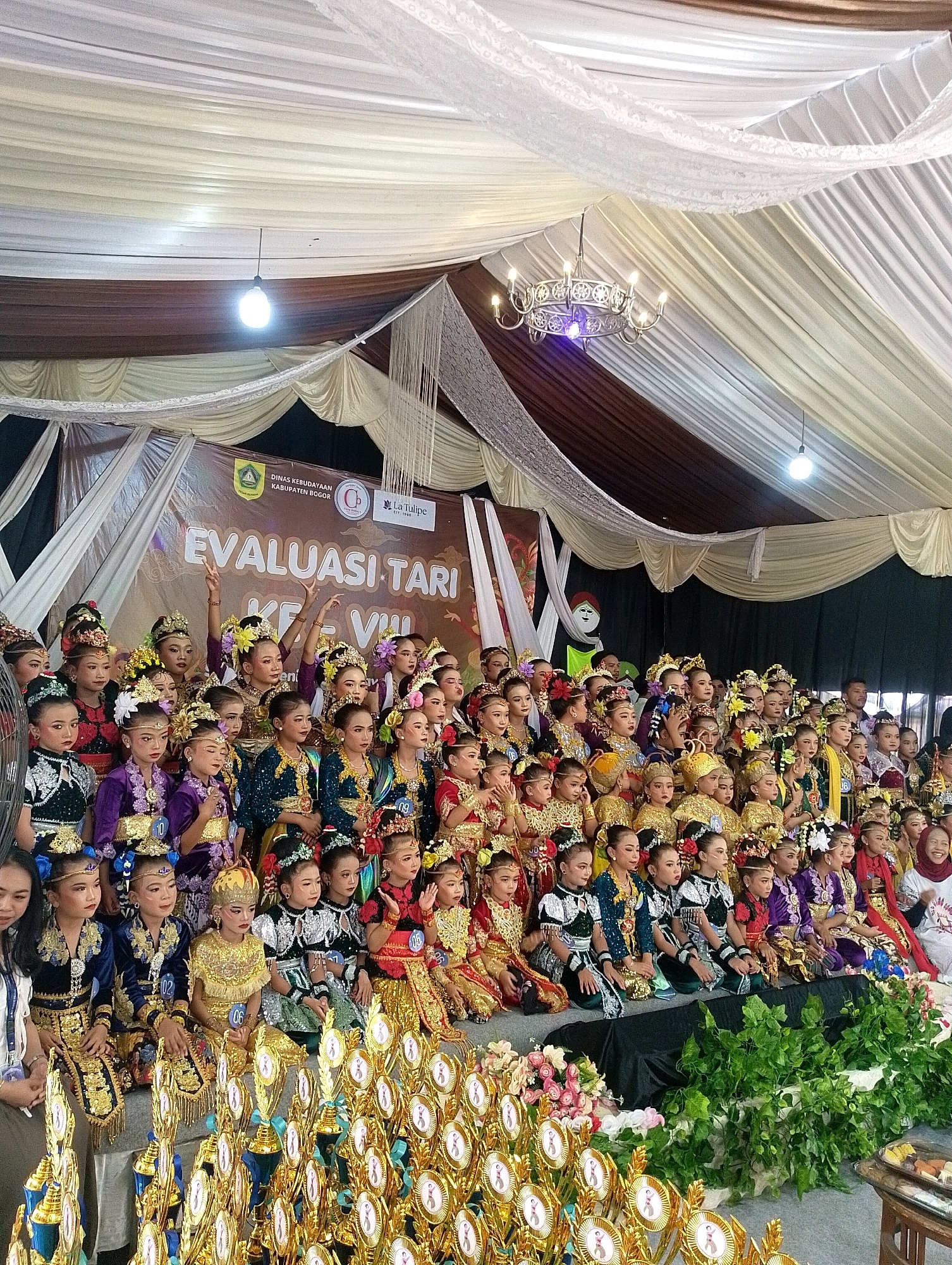 A large group of young girls, dressed in traditional Indonesian dance costumes, are posing for a photograph. The costumes are ornate, featuring gold embroidery, colorful fabrics, and elaborate headdresses. Many of the girls are wearing makeup and have their hair styled. They are seated and standing on a raised platform, with a banner in the background that reads "EVALUASI TARI KE VIII". This translates to "Dance Evaluation VIII" and suggests the event is a competition or performance. The background is decorated with draped fabric in shades of white and brown, and a chandelier hangs from the ceiling. In front of the girls are numerous trophies, indicating an awards ceremony or competition. The scene appears to be indoors, possibly in an event hall or community center in Gunung Putri, Indonesia. The overall atmosphere is festive and celebratory, with the girls looking proud and excited.