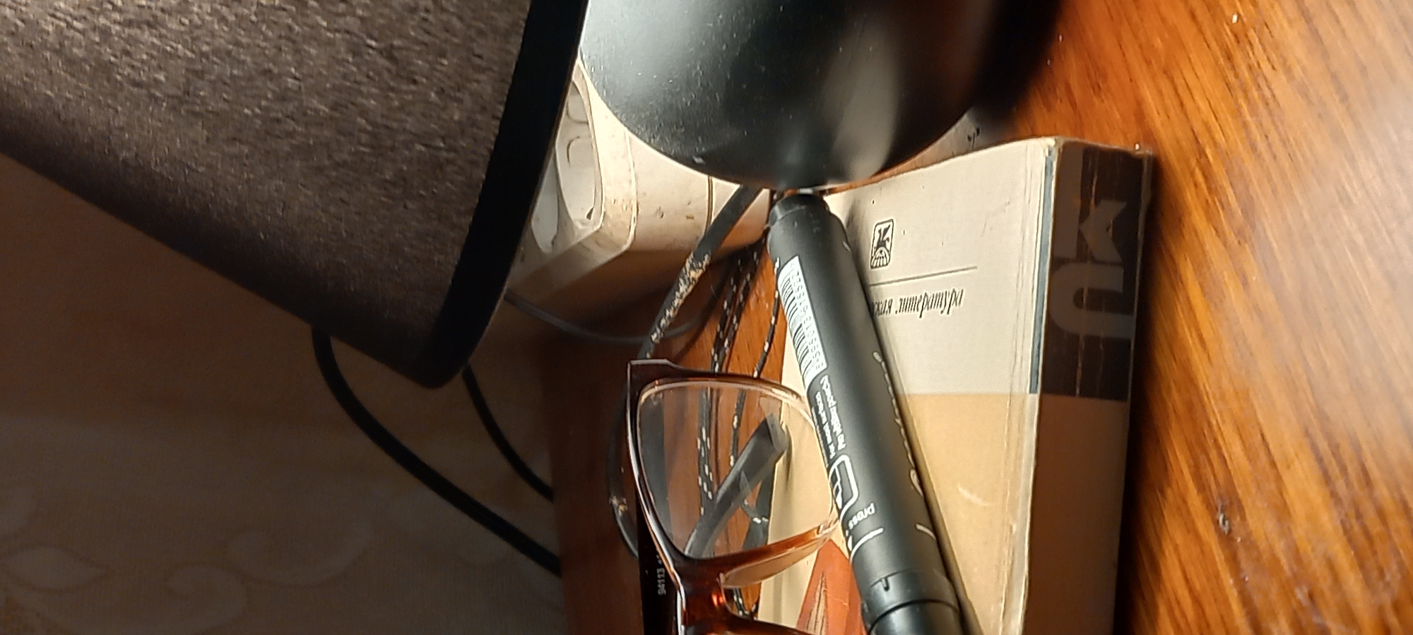 The image captures a close-up, dimly lit, horizontal view of objects arranged on a wooden surface, likely a bedside table, in Kryvyi Rih, Ukraine. A black lamp, with a textured fabric shade on the left and its base at the top center, serves as the primary light source, suggesting an evening or nighttime setting. Behind the lamp's base, a white electrical outlet and connected wires are visible.
A pair of brown-framed eyeglasses rests on the surface, with the number "94113" visible on one arm. A black marker or pen lies across the eyeglasses and a light-colored book. The marker displays partial text including "Press" and what appears to be a numerical value like "0.6" or "0.8" on a white label.
The book has a brown spine featuring a dark, stylized logo, possibly depicting 'K' and 'U'. Its cover shows Cyrillic text, partially discernible as "ская литература" (skaya literatura), meaning "literature," along with a distinct square logo containing an abstract design. The wooden surface beneath the items exhibits a visible grain and a subtle, embossed decorative pattern in the lower left area. No people are present in the scene.
