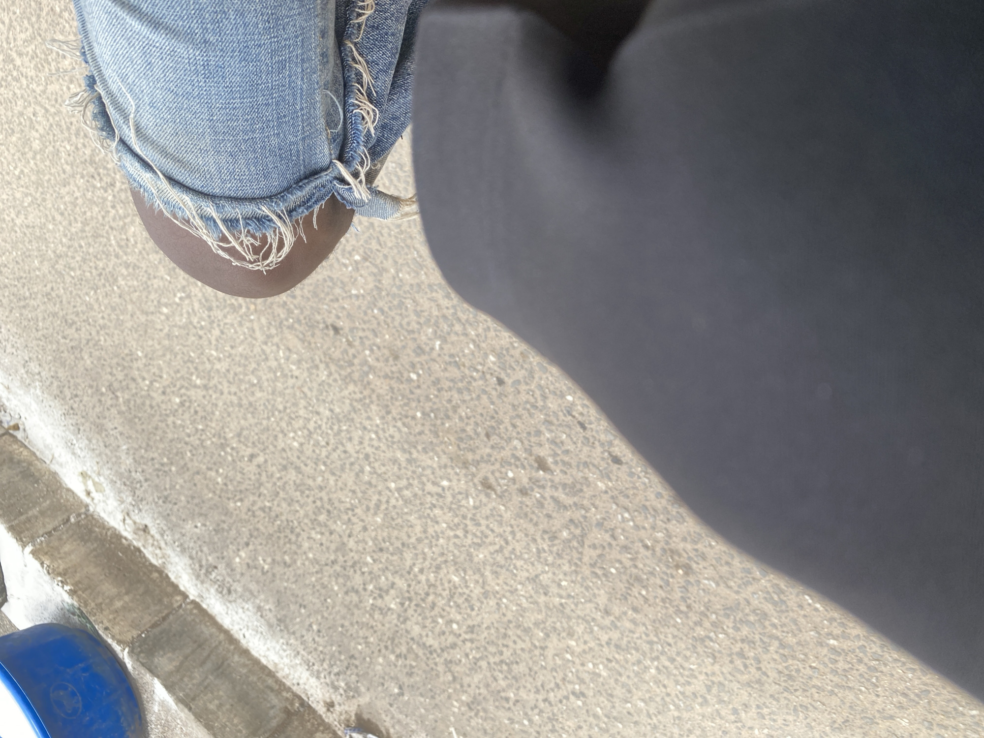 The image shows a close-up of a person's knee and lower leg, wearing distressed denim shorts and dark leggings. The person is standing on a rough, asphalt-like surface, possibly a sidewalk or road, with a concrete curb visible in the lower left corner. A blue plastic object, possibly a bucket or container, is partially visible at the very bottom edge of the frame. The lighting suggests it is daytime, with the sun casting a shadow. The scene appears to be outdoors in an urban or semi-urban environment, given the pavement and curb. The distressed nature of the denim, with frayed edges, is a notable detail. There is no visible text in the image. The overall impression is a snapshot of a person's lower body in an everyday setting.