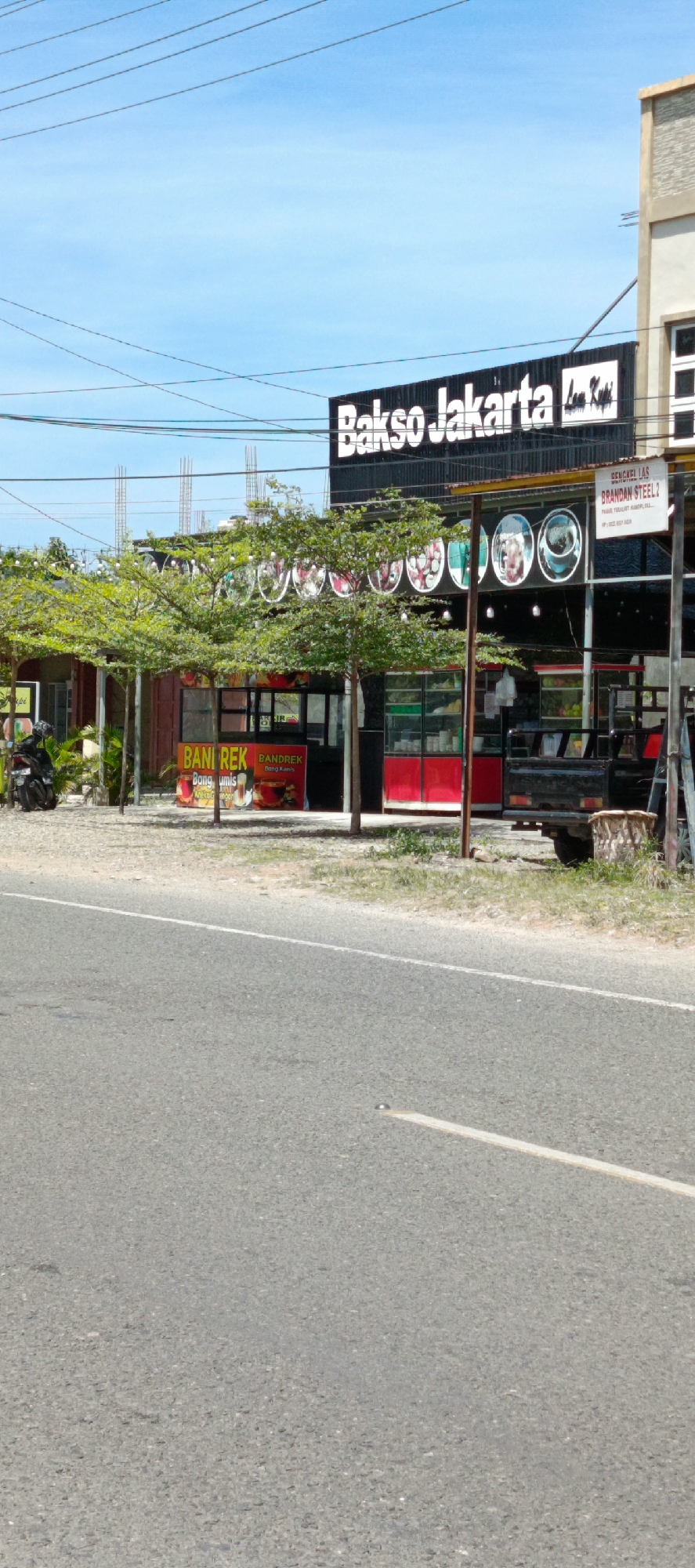 This is a full-frame, eye-level, outdoor, vertical shot of a commercial establishment in the city of Lampisang, Indonesia. The establishment, identified by a large black sign with white lettering reading "Bakso Jakarta," appears to be a restaurant or food stall. In front of the establishment are several trees with lush green foliage and strings of small lights. A portion of a building with a stone-like facade is visible on the right.
A small kiosk or counter with red and white signage is positioned in front of the main establishment. The signage on this kiosk reads "BANI REK" and "BANDREK" with additional text in Indonesian script. A motorcycle is parked to the left of the kiosk, and a small truck is parked to the right. The ground in front of the building is a rough, sandy surface with some scattered vegetation. A paved road with a white line marking runs across the foreground of the image. The sky above is bright blue with scattered clouds. The image is captured on a sunny day.