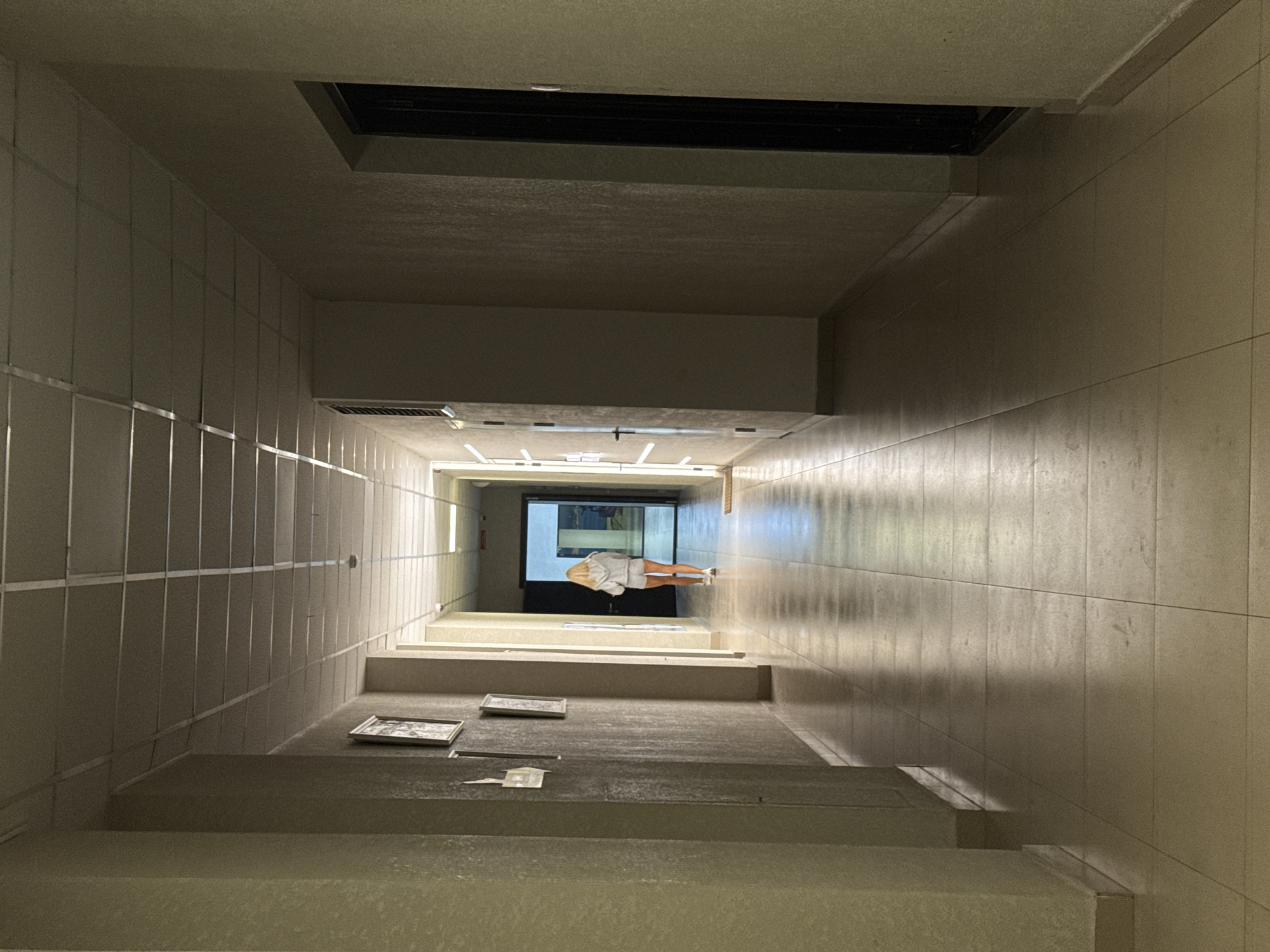 A blonde woman wearing a grey hoodie and shorts walks down a tiled hallway in a building. The hallway has beige walls and a white drop ceiling with fluorescent lighting. There are framed pictures on the left wall. The hallway extends into the distance, where there is an open doorway with light coming through. The floor is made of large square beige tiles that reflect the light. The scene is illuminated by the overhead lights, casting shadows down the hallway.