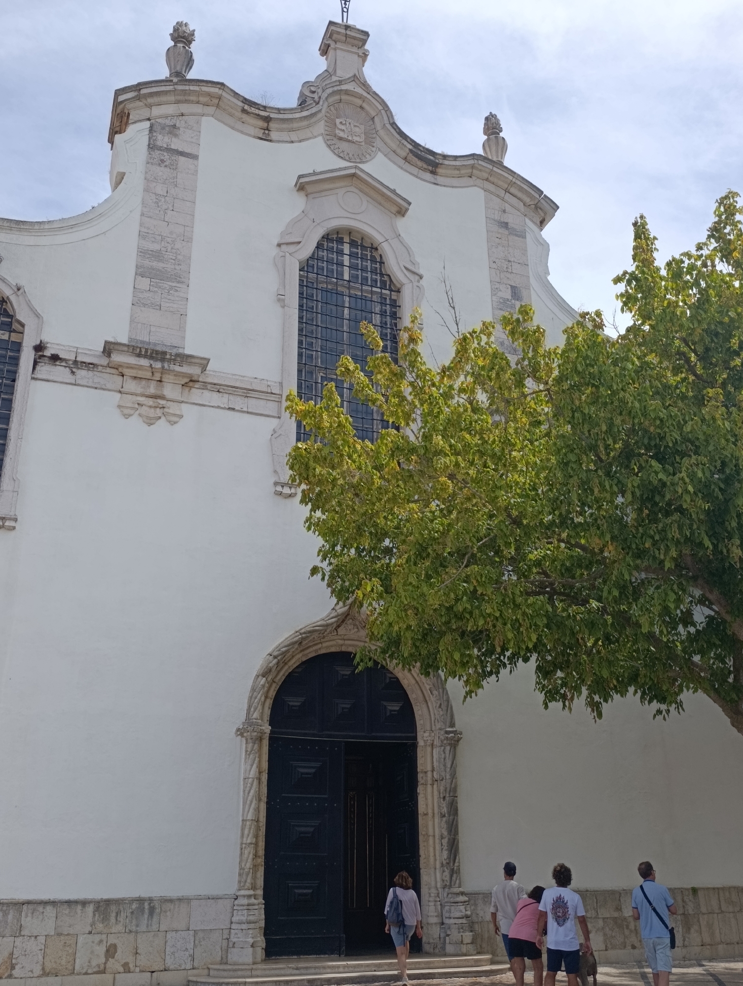 A white church facade with a large, dark wooden door framed by an ornate stone archway dominates the center of the image. A mature tree with vibrant green leaves extends from the right side, partially obscuring the upper right portion of the church. Several people, viewed from behind, are walking towards the church entrance, suggesting they are entering or approaching. The architecture appears to be historical, with stone accents and decorative elements on the facade, indicative of a location in Setúbal, Portugal. The sky is a pale blue with some clouds, suggesting daytime.