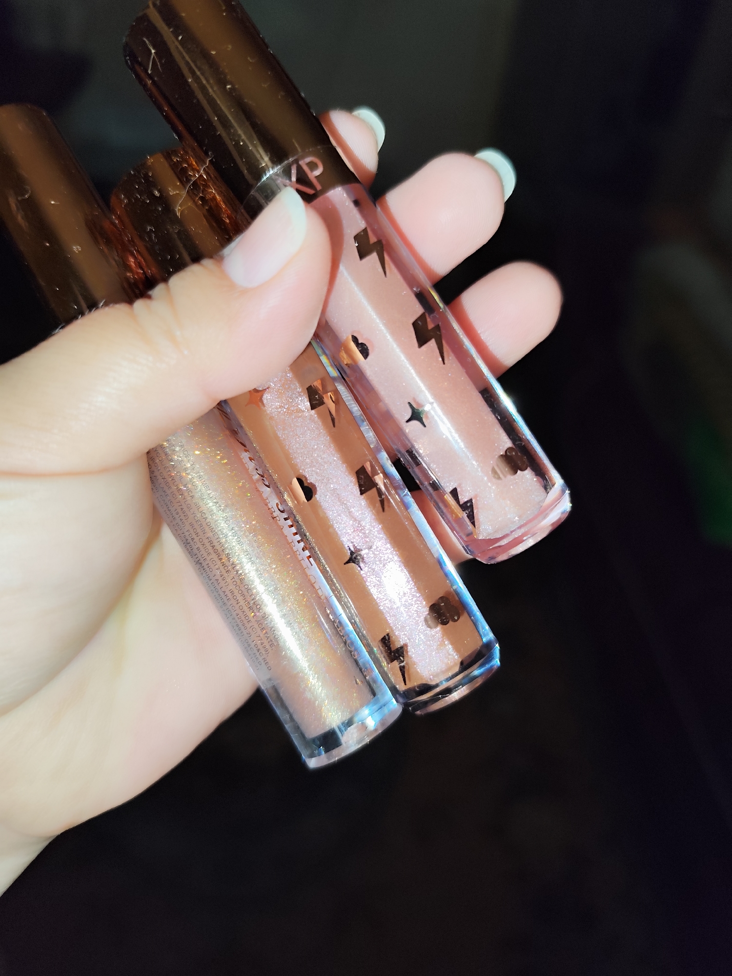 A hand holds three lip gloss tubes. The tubes are clear with bronze lids and contain a shimmery liquid. The lip gloss tubes have small black graphic designs. The hand is fair-skinned with light pink nail beds and white-tipped nails. The background is out of focus and dark. There is no indication of the city of Sarıyer, Turkey.