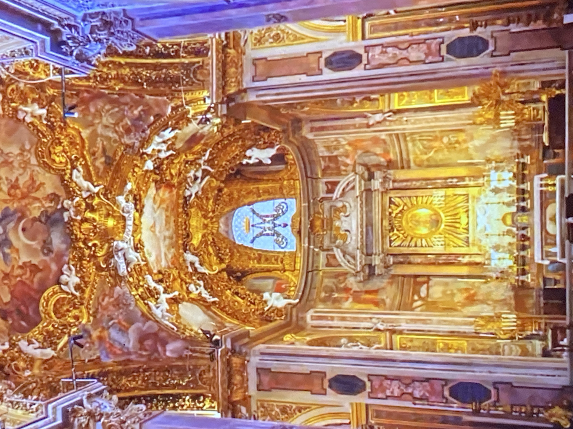 The image captures the ornate ceiling of a building, likely a church or cathedral, in Tinaquillo, Venezuela. The architectural style is baroque, heavily adorned with gold leaf, sculptures of angels, and intricate paintings. The ceiling design features a central oval window with blue stained glass. A radiant sun motif is prominently displayed, possibly representing a religious symbol. The overall scene conveys a sense of grandeur and religious significance.