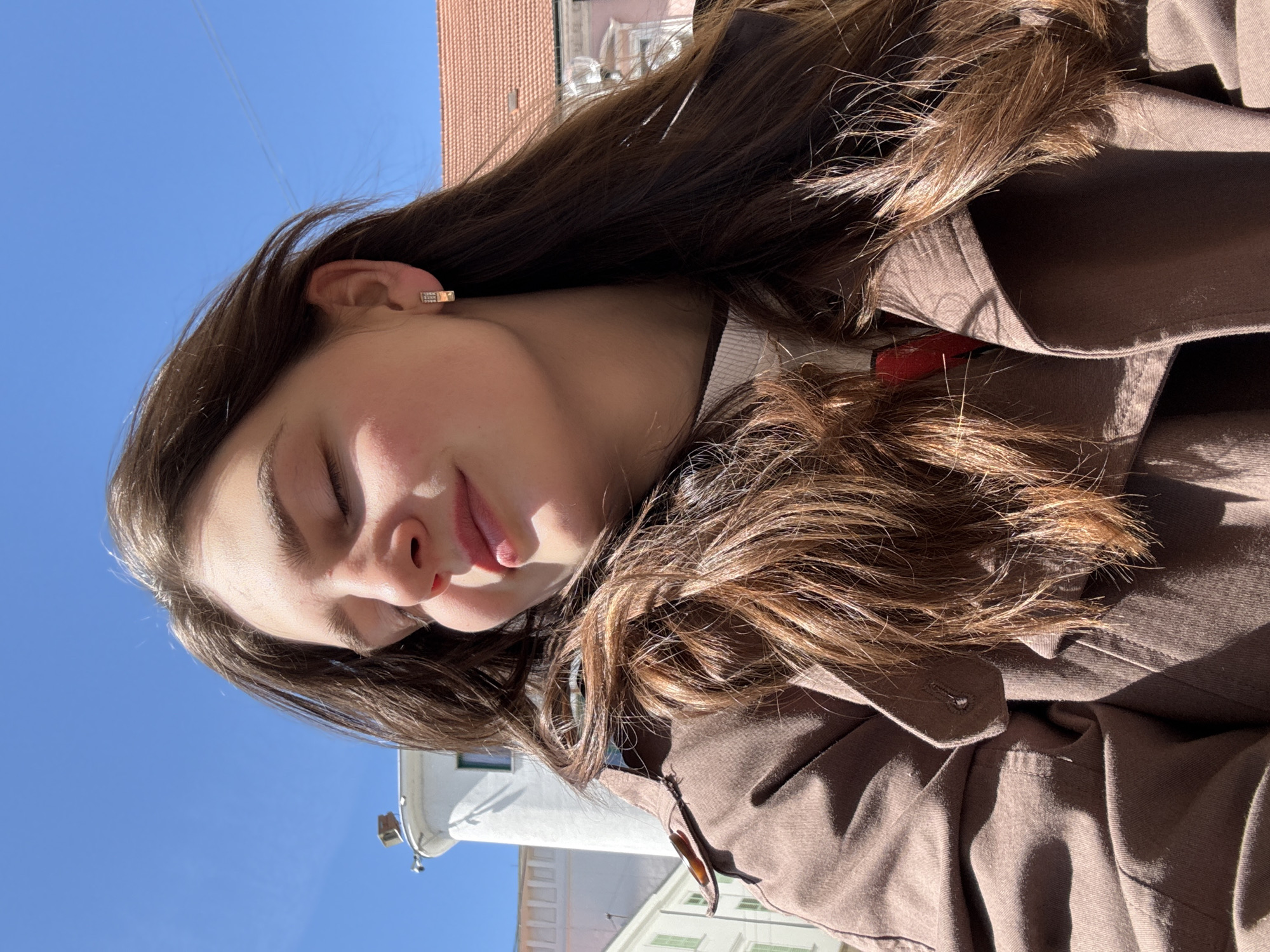 A young woman with long brown hair is outdoors, eyes closed, basking in the sunlight. She is wearing a brown coat and a light-colored top underneath. Her face is angled towards the sun, and her lips are slightly parted. There are buildings in the background, one with a reddish-brown roof and another white building with green shutters and what appears to be a clock tower. The blue sky is visible in the upper left corner. Based on the architecture, the scene appears to be in the city of Trnava, Slovakia.