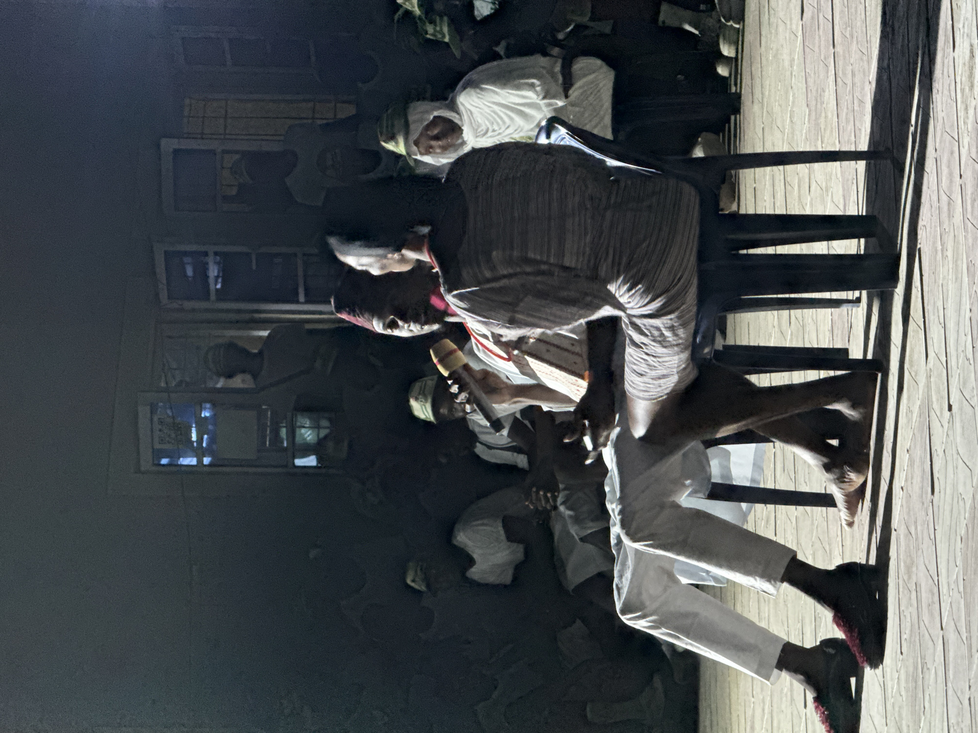In Maiduguri, Nigeria, an assembly of people is seated on chairs. One man is standing, holding a microphone. The individuals are dressed in a mix of casual wear and traditional white attire with head coverings. The setting appears to be near a building with windows.