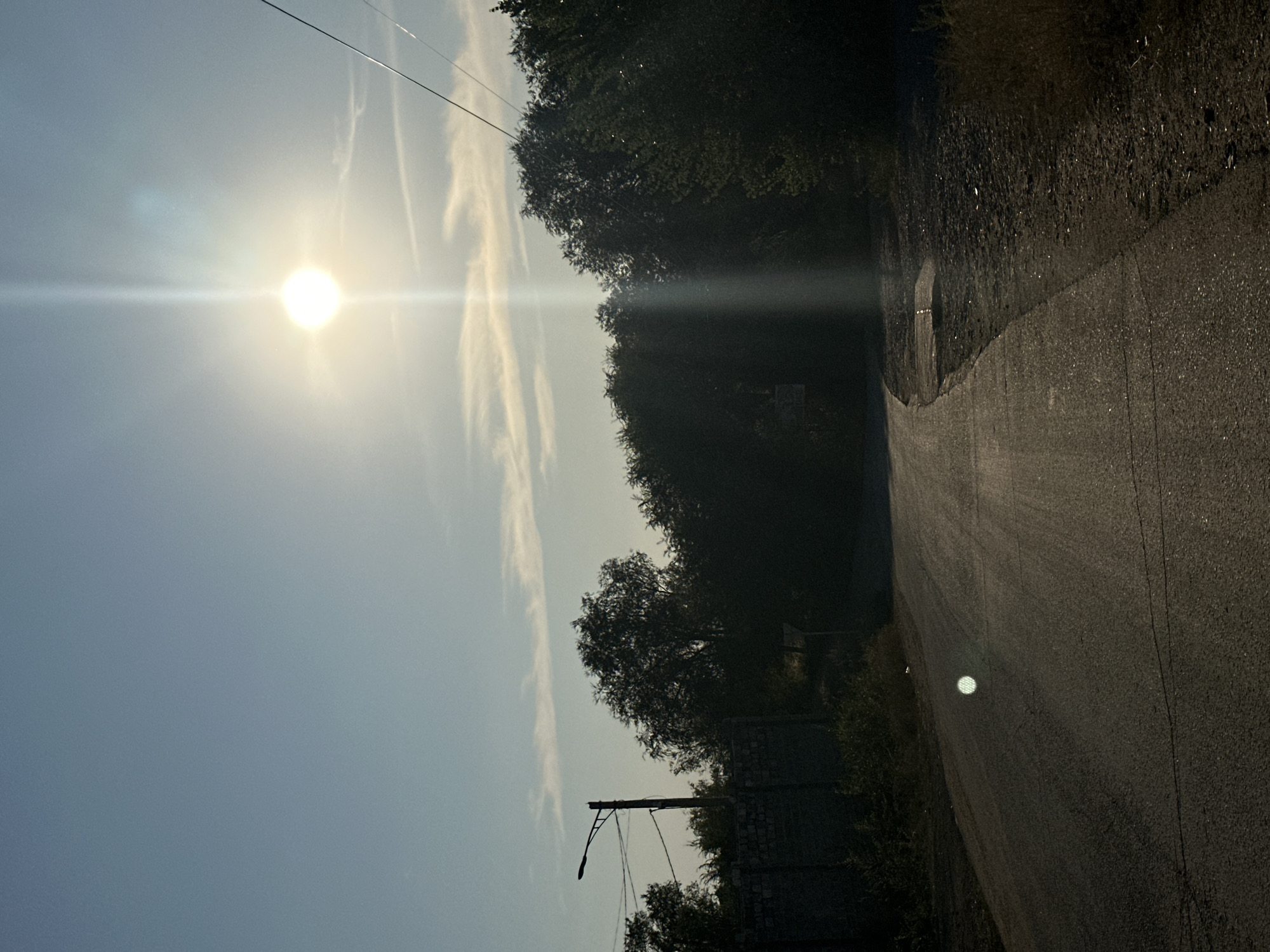 The image depicts a rural road in Energetik, Russia, bathed in the bright, low-lying sun. The road curves gently to the left, its asphalt surface showing signs of wear and tear. To the left of the road, a stone wall is visible, topped with sparse vegetation. A lamppost with wires attached stands to the left of the wall. A dense line of trees forms a dark silhouette against the sky, bordering the right side of the road. The sky is a pale blue with wispy clouds and a strong sunbeam piercing through, creating lens flare. The overall impression is one of a quiet, somewhat isolated setting.