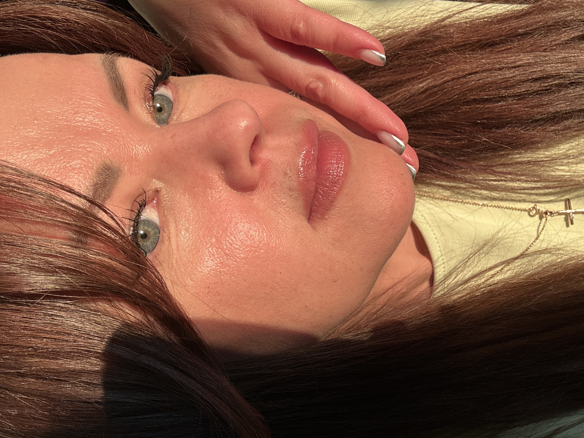 This close-up, vertically oriented portrait features a woman with long, dark brown hair, lying down with her head turned upwards. Her face, including her grey-blue eyes, nose, and glossy pink lips, is clearly visible. Strong, directional sunlight illuminates the right side of her face and hair, creating pronounced highlights on her skin and a natural, dewy appearance. Her dark brown hair, with visible reddish undertones, is spread out around her head and shoulders, shining where it catches the light.
One of her hands, with manicured nails featuring silver-toned French tips, is positioned gently on her hair near her head. She wears a light-colored, possibly cream or pale yellow, garment and a delicate gold chain necklace with a small cross pendant visible on her chest.
The strong, warm lighting suggests a bright, sunny daytime environment, possibly early morning or late afternoon, creating a serene and calm mood. The woman's expression is neutral and contemplative. There is no visible text within the image. While no specific architectural or landscape elements are present, the context indicates the scene is set in Chaikovsky, Russia.