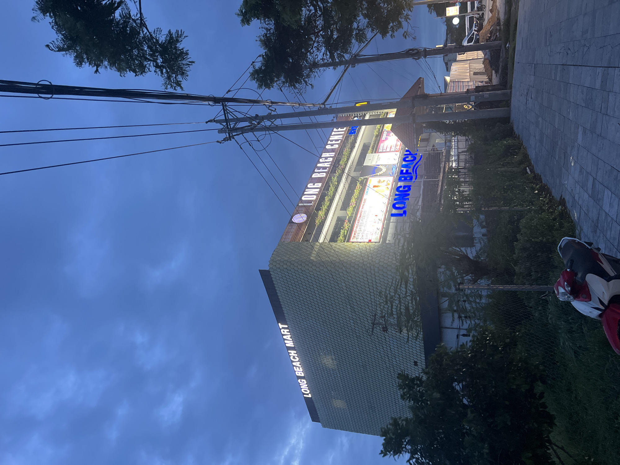 The image shows the exterior of a multi-story building identified by illuminated signs as "LONG BEACH MART" and "LONG BEACH CENTER" in Phú Quốc, Vietnam. The building has a distinctive facade with a textured, scale-like pattern on its upper levels and illuminated signage on its roof and front. Greenery is visible along the lower levels of the building, and a balcony area with plants is also present.
In the foreground, a red and white motorcycle is parked on a paved walkway. The walkway is constructed of grey interlocking pavers. To the right of the walkway and in front of the building, there is a sidewalk and then a street with power lines and utility poles overhead. Further down the street, construction materials are visible, suggesting ongoing development. The sky is overcast with a deep blue hue, indicative of dusk or a cloudy day. The overall scene depicts an urban commercial area in Phú Quốc.