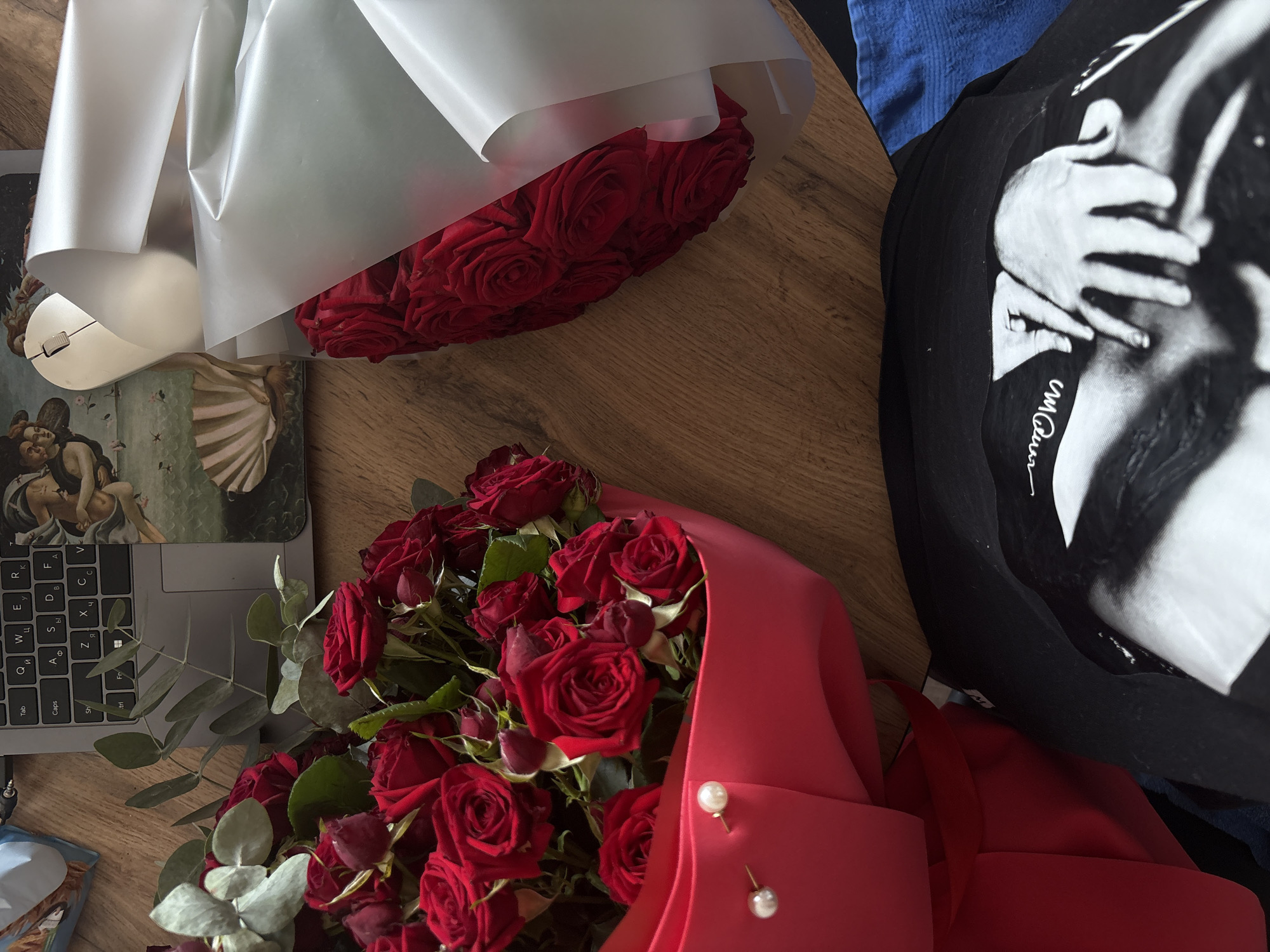 The image shows a top-down view of a wooden surface with several objects arranged on it. In the foreground, a black t-shirt with a black and white photographic print of hands is visible. To the left, a large bouquet of red roses wrapped in red paper with two pearl embellishments is arranged. To the right, another bouquet of red roses is partially wrapped in white paper. A laptop with a Russian keyboard is situated behind the red rose bouquet, and a computer mouse is placed next to it. In the background, a decorative mat with an image of "The Birth of Venus" by Sandro Botticelli is visible. A blue cloth is present in the bottom right corner of the image. The scene appears to be an indoor setting, likely a desk or table, and does not contain any specific geographical markers to indicate its location in Rostov-on-Don, Russia.