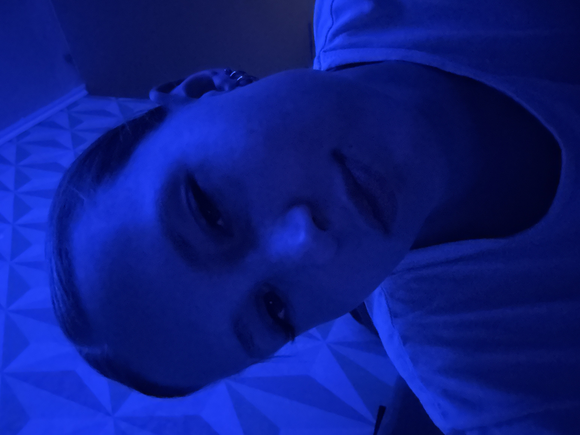 A close-up shot of a person's face illuminated by a strong blue light, possibly a neon light or LED lighting, indoors. The person appears to be wearing a light-colored top, possibly a white t-shirt. The face is partially obscured by shadows. The background features a geometric pattern, possibly a textured wall or paneling. The image does not provide enough information to identify a specific location in Chaikovsky, Russia. It could be taken anywhere indoors with similar lighting conditions.