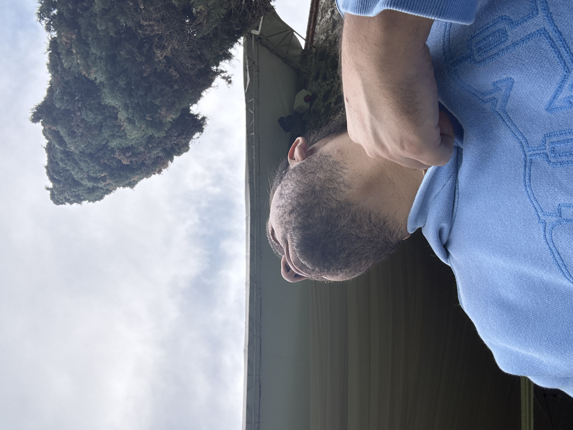 A low-angle shot shows a man in a light blue sweatshirt standing outdoors in Santa Ana Acozautla, Mexico. A tree is visible in the upper left, and a building or structure with a grey roof is above the man. The sky is cloudy. The man is adjusting the collar of his sweatshirt with his right hand. He has dark hair and a beard.