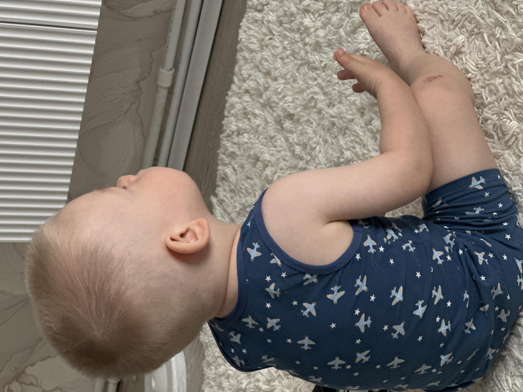 A young child wearing blue pajamas with white airplane and star patterns lies on a light-colored shag rug indoors. The child's head is near a set of closed white blinds. The child has blonde hair and a scrape on one knee. The location appears to be a residential setting.