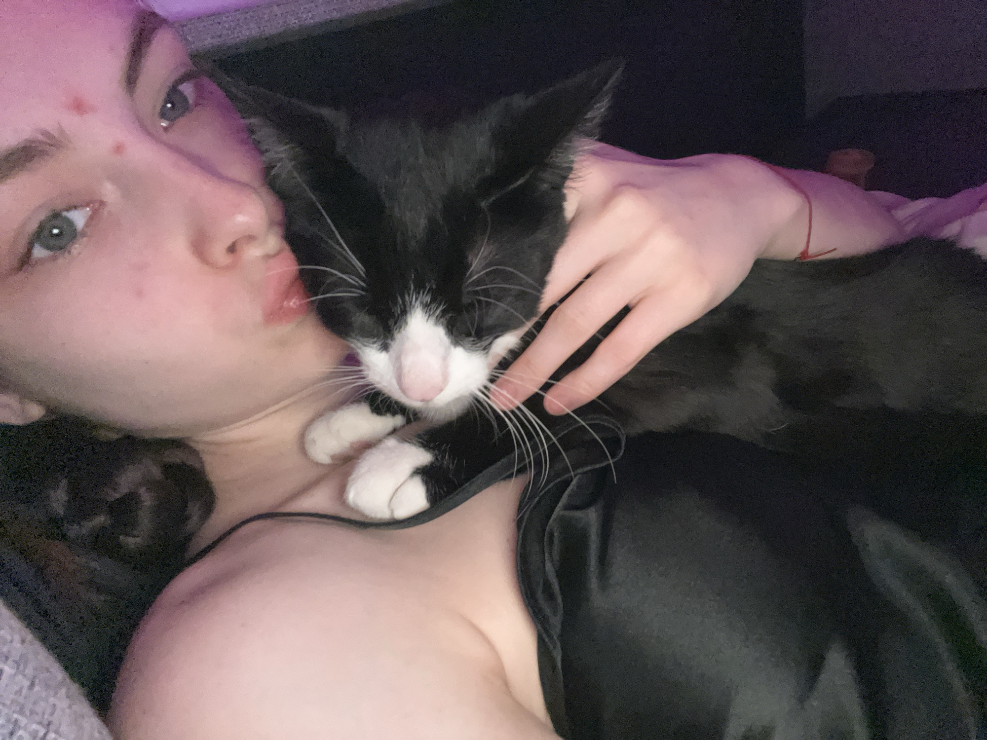 A young woman with fair skin, hazel eyes, and light brown hair lies on a soft, gray-toned surface. She has a few blemishes on her forehead. She is close to a black and white cat, possibly kissing it. The cat has a white nose, white paws, and closed eyes. The woman's right hand is gently holding the cat. She is wearing a black, silky top with thin straps. A red string bracelet is visible on her wrist in the background. The location and city are not discernible from the image.