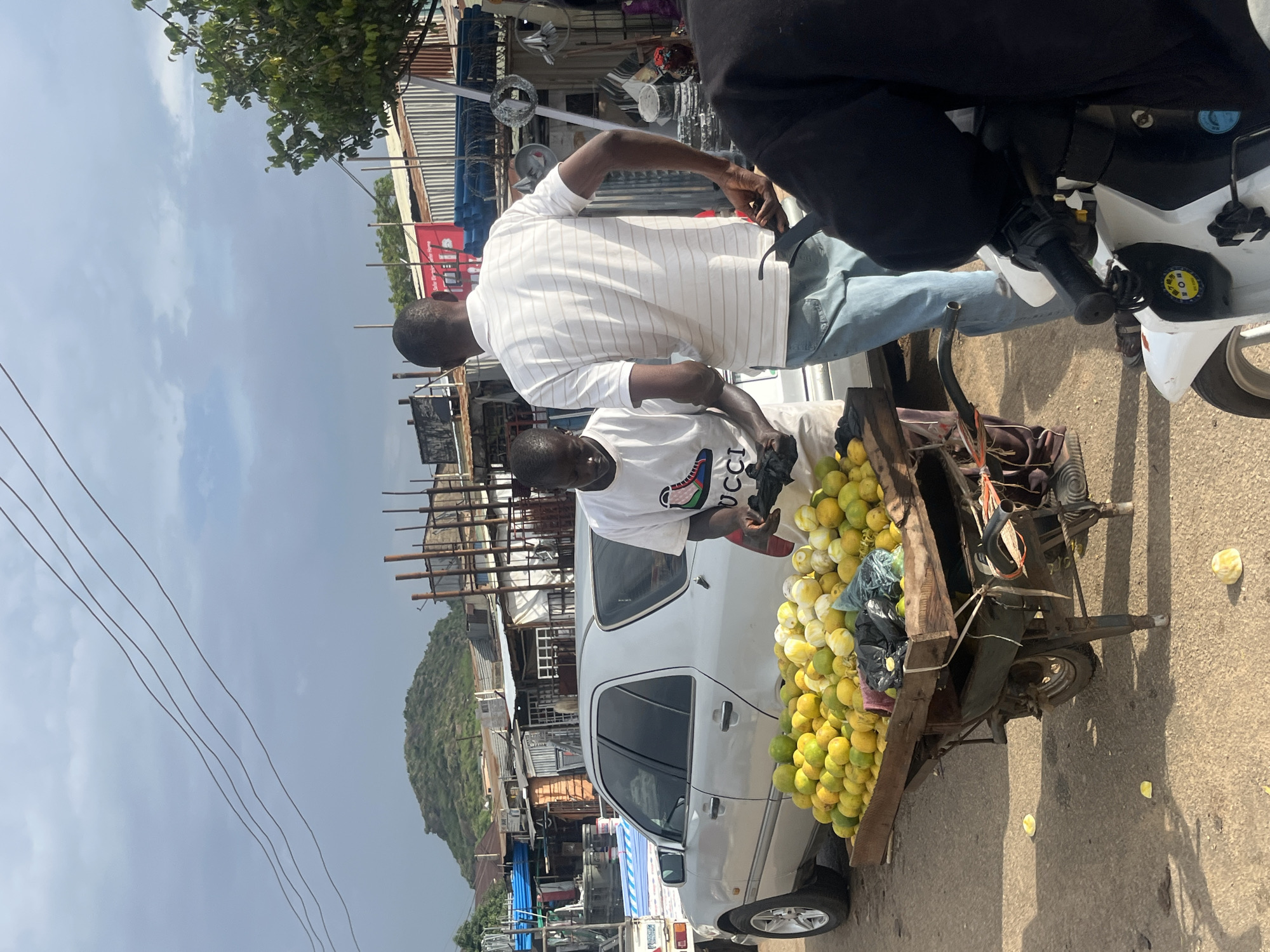Two men are engaged in a transaction near a fruit cart laden with lemons and limes on a dusty street. A silver sedan is parked behind the cart, and further back, various structures with corrugated metal roofs and exposed scaffolding are visible. In the distance, a green, rocky hill rises against a cloudy sky. A motorcycle is parked in the foreground on the right. The scene appears to be in a commercial or market area.