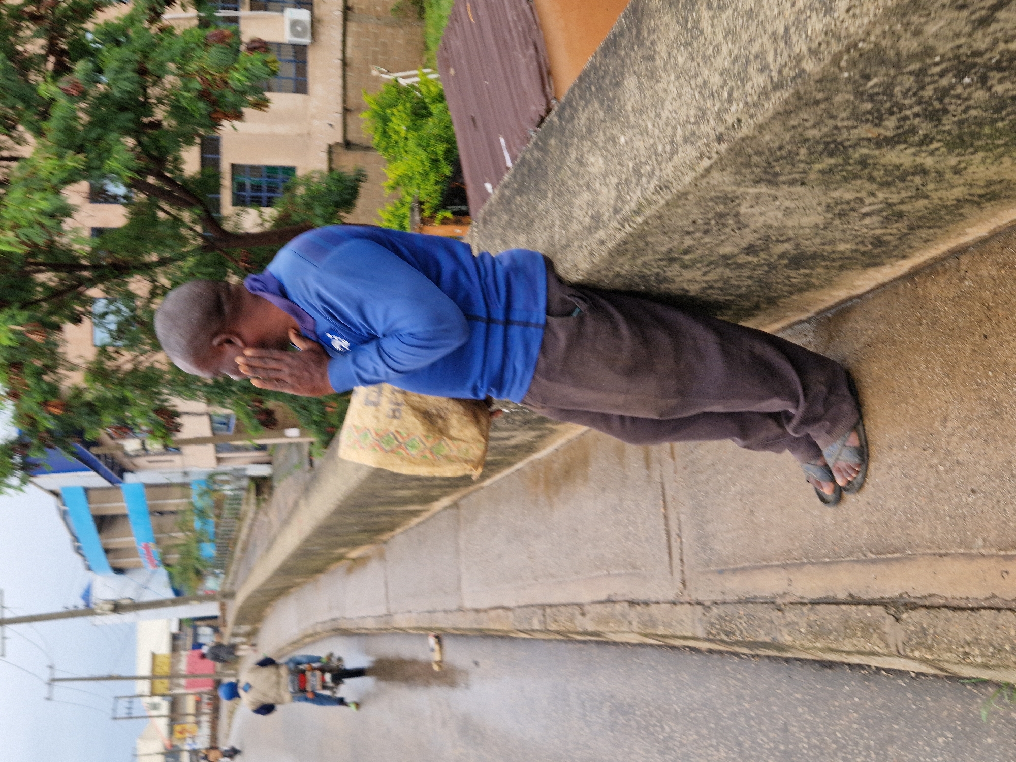A man with dark skin, bald head, and a blue long-sleeved shirt is leaning on a concrete wall, holding a light brown bag with both hands to his face. He is wearing dark pants and dark sandals. He is standing on a sidewalk next to a road. The road is wet, suggesting it might have rained. A person on a motorcycle is visible on the road in the background, heading away from the camera. The surrounding environment appears to be urban, with buildings and trees visible. The man's position and the bag held to his face might suggest he is experiencing discomfort or grief.